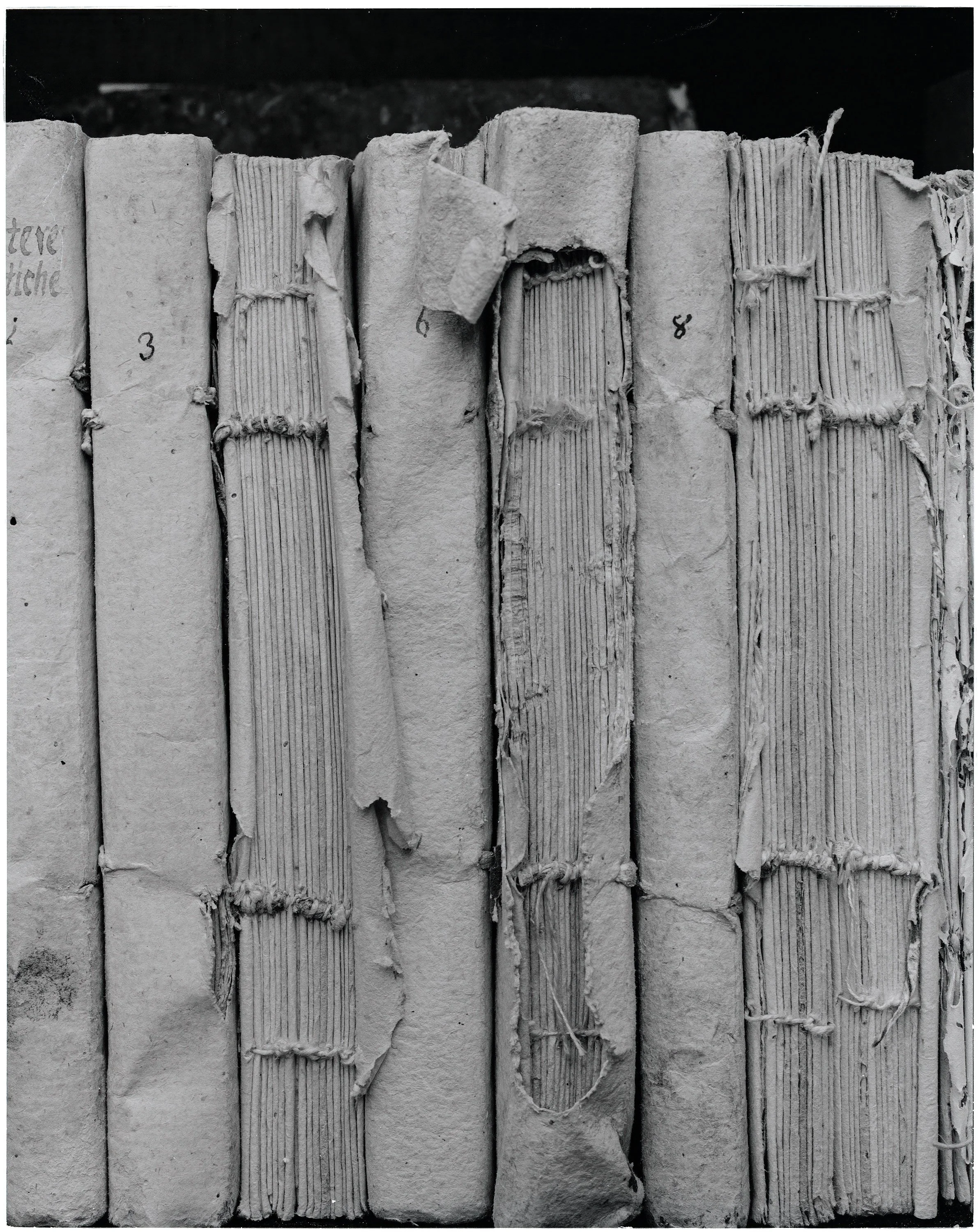 Close-up black and white photograph of old, worn books with visible pages and bookmarks, arranged upright on a shelf.