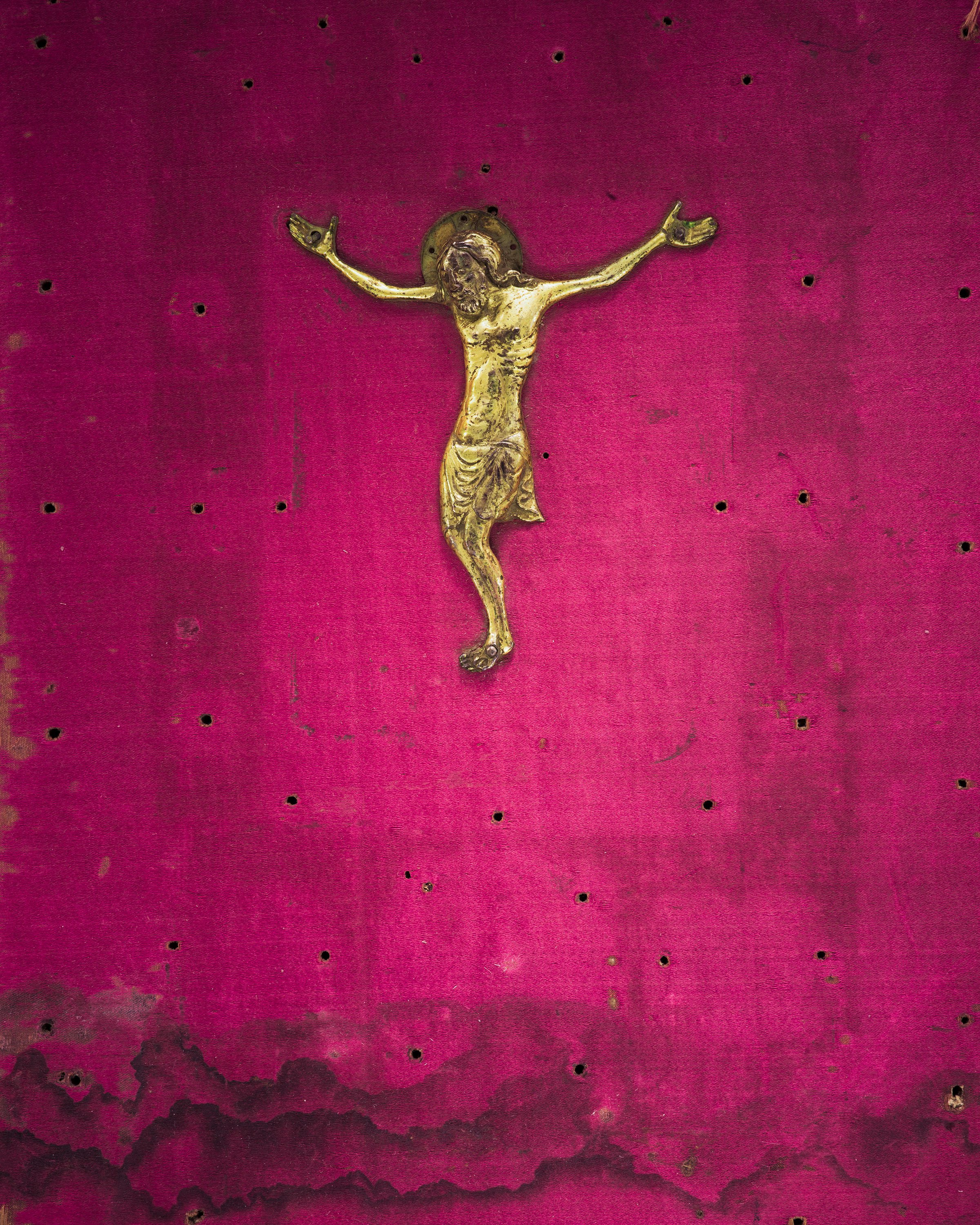 A metal crucifix depicting Jesus Christ on a pink wall with small, evenly spaced holes and scattered dark spots.