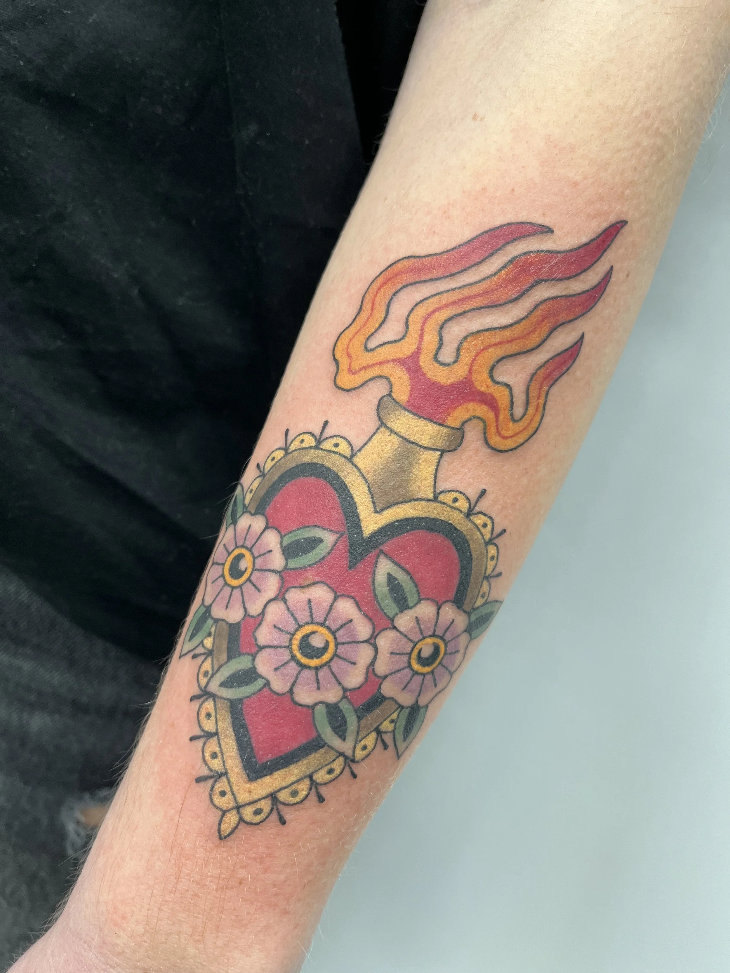 Tatouage d'un cœur avec des fleurs roses et un motif représentant une flamme sur l’avant-bras.