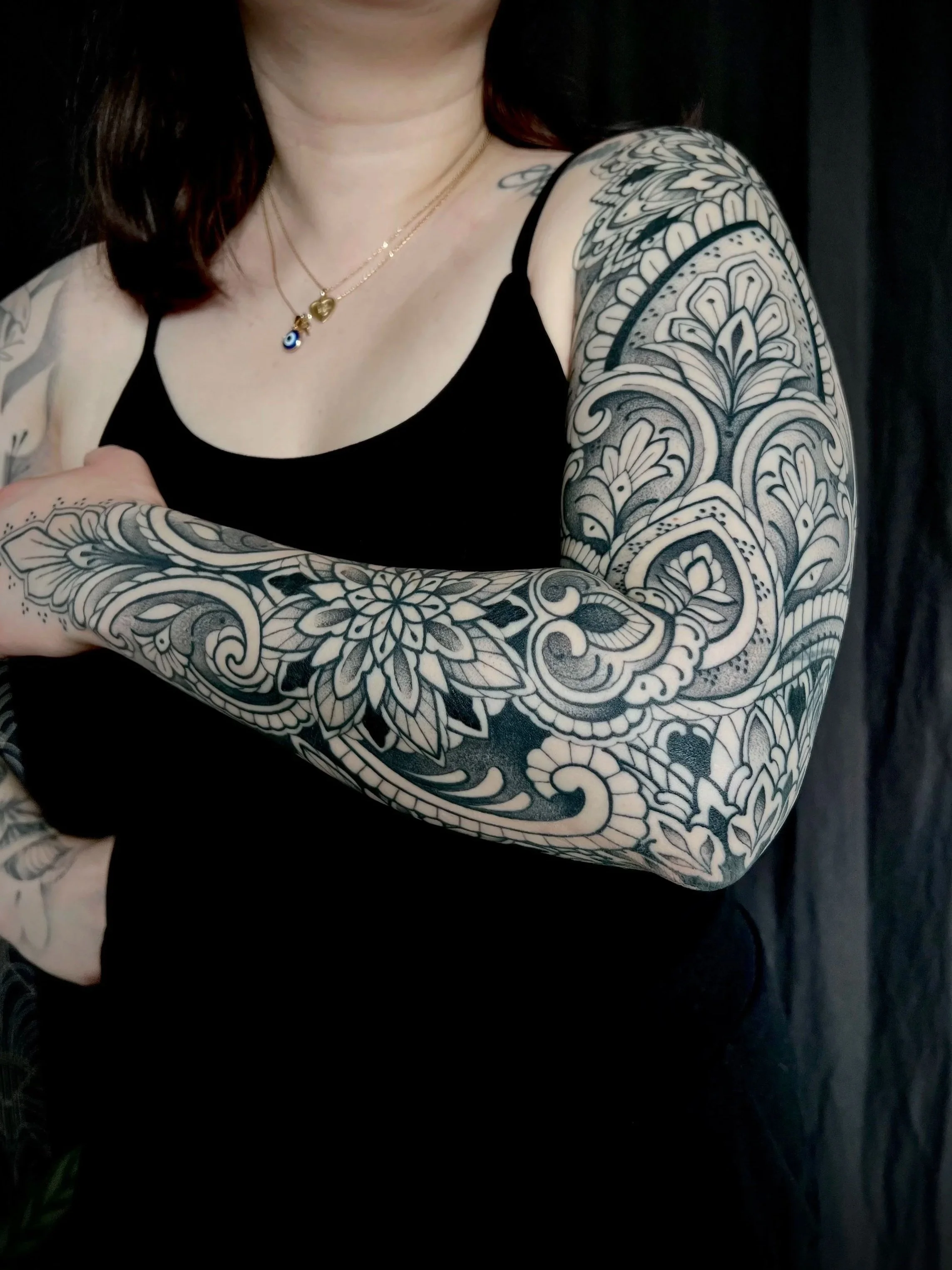Une femme porte un t-shirt noir et exhibe des tatouages ornementaux complexes et détaillés sur le bras et l'épaule.
