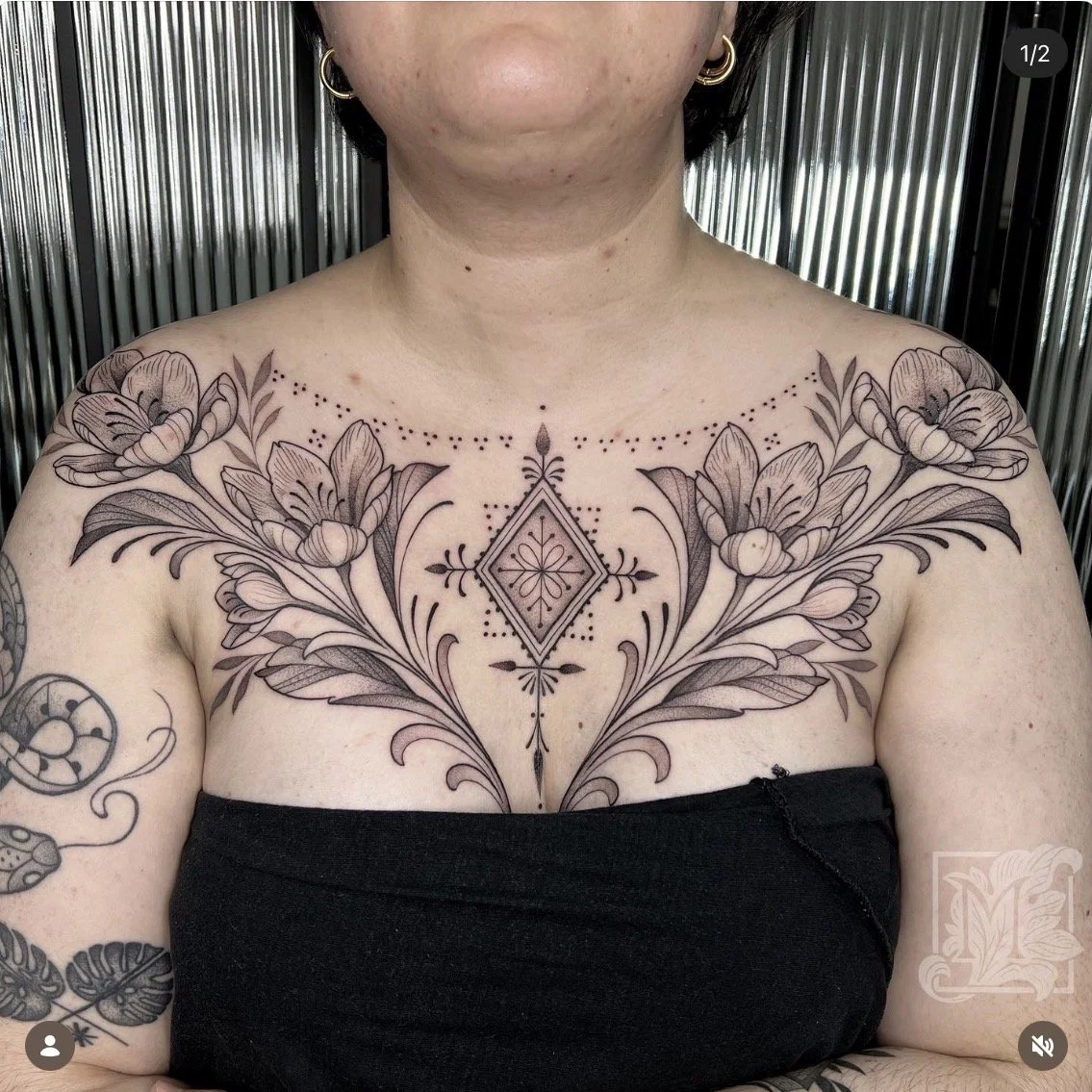 Tatouage floral sur la poitrine d'une femme, avec des motifs de fleurs, de feuilles, et un motif d’inspiration berbère au centre, en noir et gris.