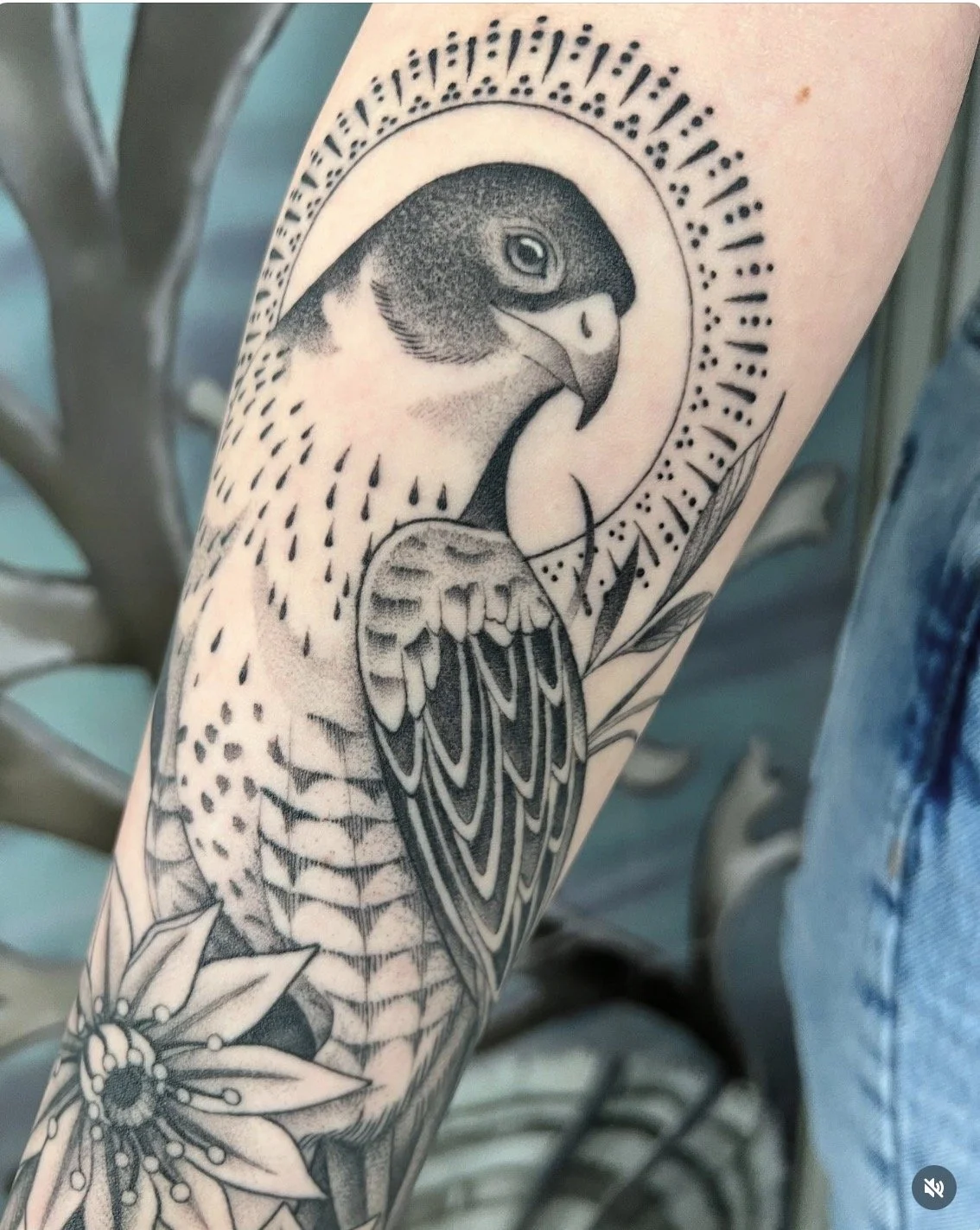 Tatouage noir et blanc d'un faucon encadré de fleurs avec un halo ornemental.