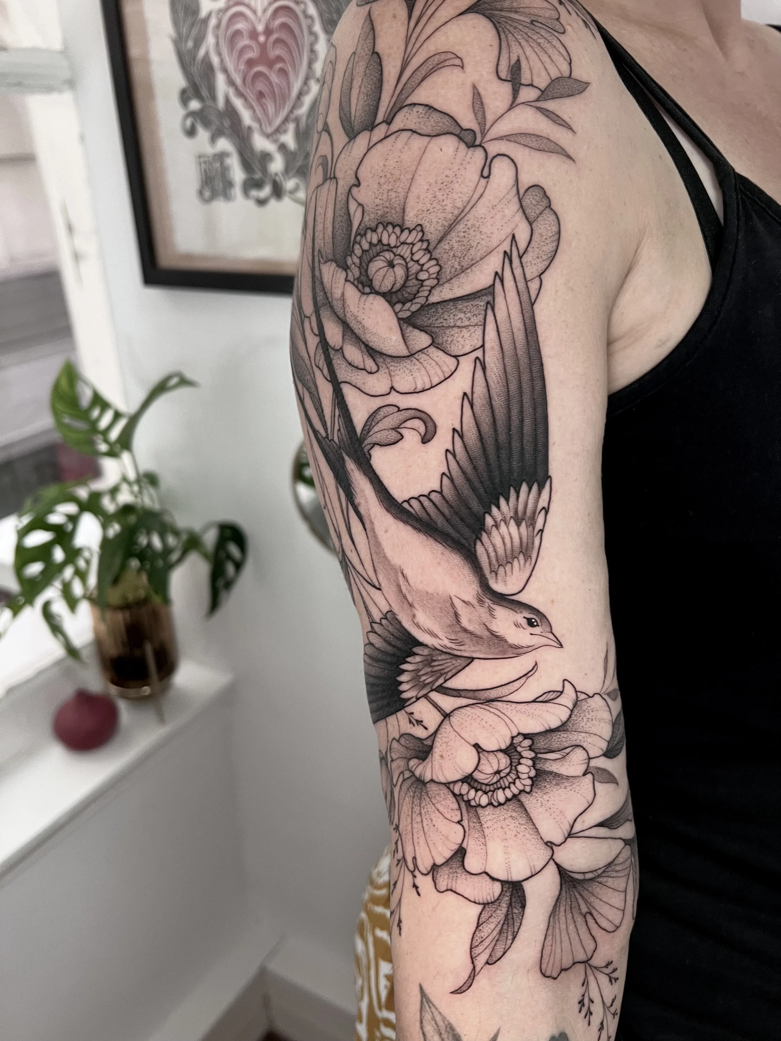 Un bras d'une personne avec un tatouage en noir et gris représentant un oiseau en vol entouré de fleurs et de feuilles.