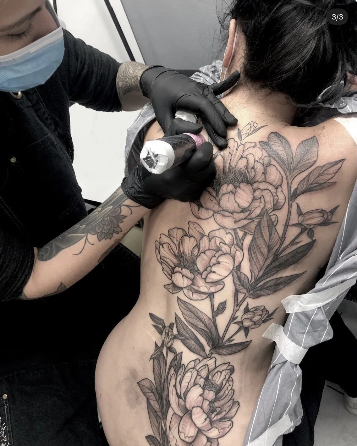 Une femme se fait tatouer un grand motif floral noir sur le dos par un tatoueur portant un masque et des gants noirs.