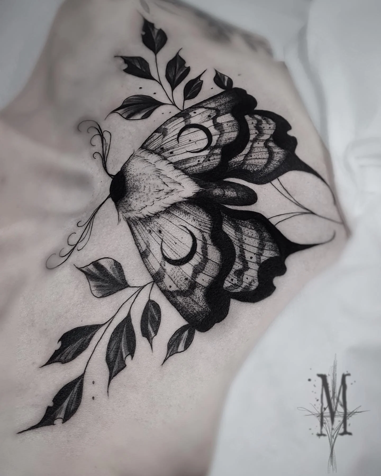 Tatouage noir d'un papillon de nuit sur la poitrine entouré de branches avec des feuilles en noir et gris.