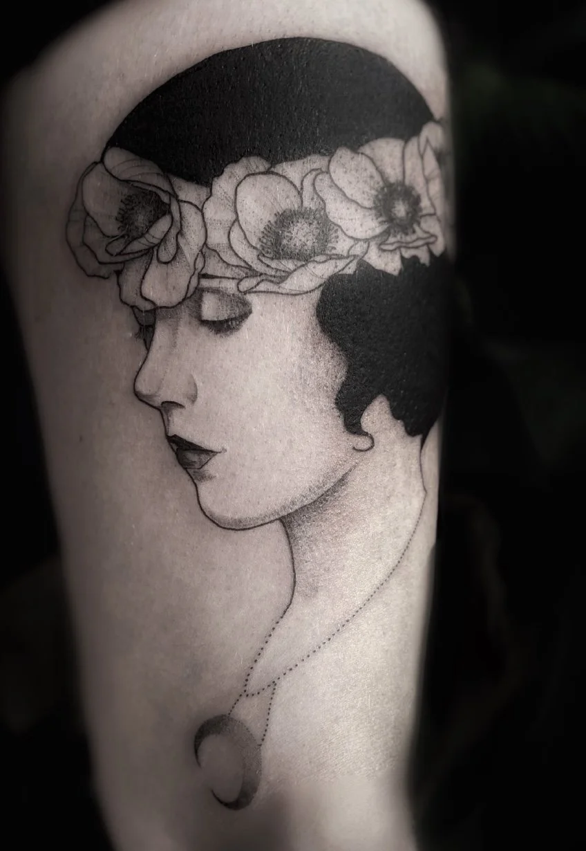 Tatouage noir et blanc d'une femme avec une couronne de fleurs sur la tête, le visage en profil, portant un collier avec un croissant et une lune.