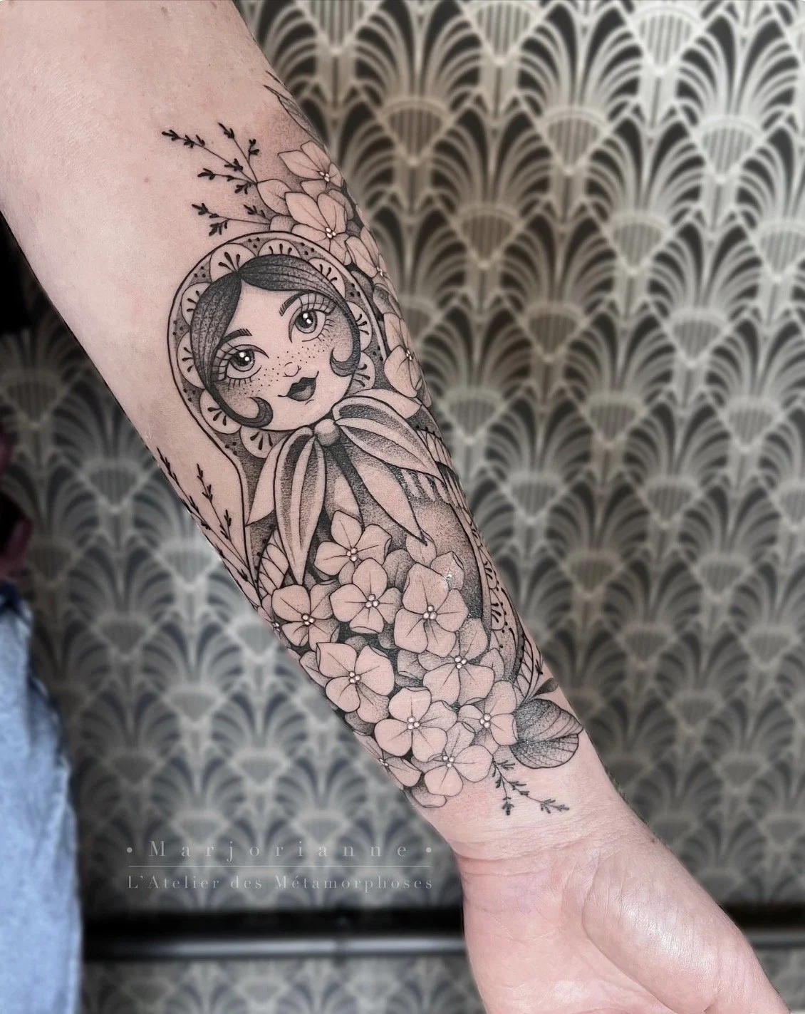 Tatouage en noir et blanc d'une magriochka avec de grands yeux, des cheveux courts et ondulés, portant un foulard ou un ruban, entourée de fleurs et de branches