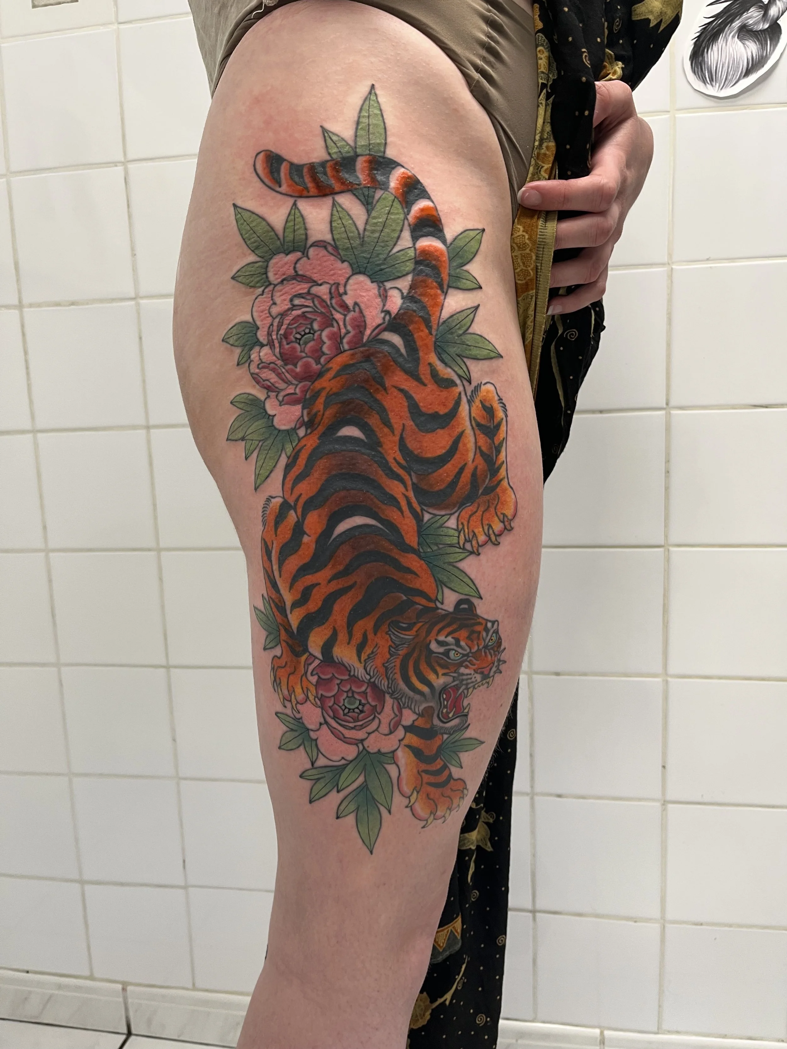 Tatouage d'un tigre orange et noir entouré de fleurs roses et de feuilles vertes, situé sur la cuisse d'une personne.