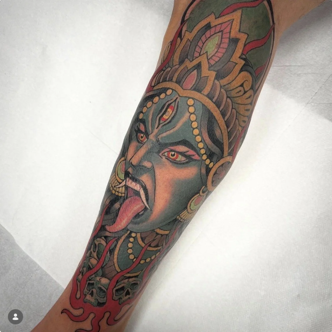 Tatouage d'une tête de la déesse Kali avec une langue rouge, portant un casque orné de perles et de motifs colorés, avec des yeux rouges et un visage expressive.