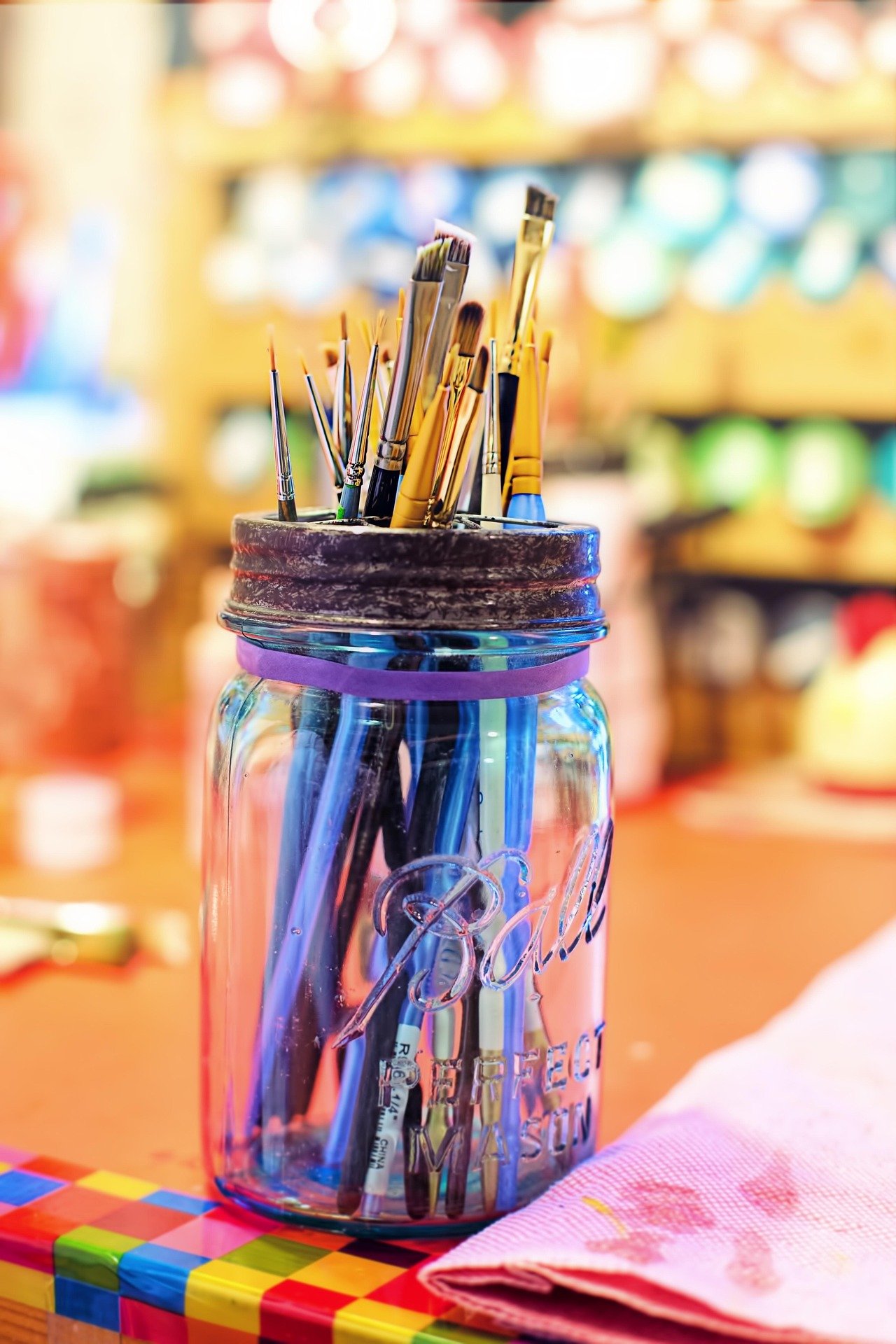 Un pot en verre contenant des pinceaux de différentes tailles et formes, placé sur une table avec un napperon rose à côté. En arrière-plan, un mur avec des bocaux colorés flous.