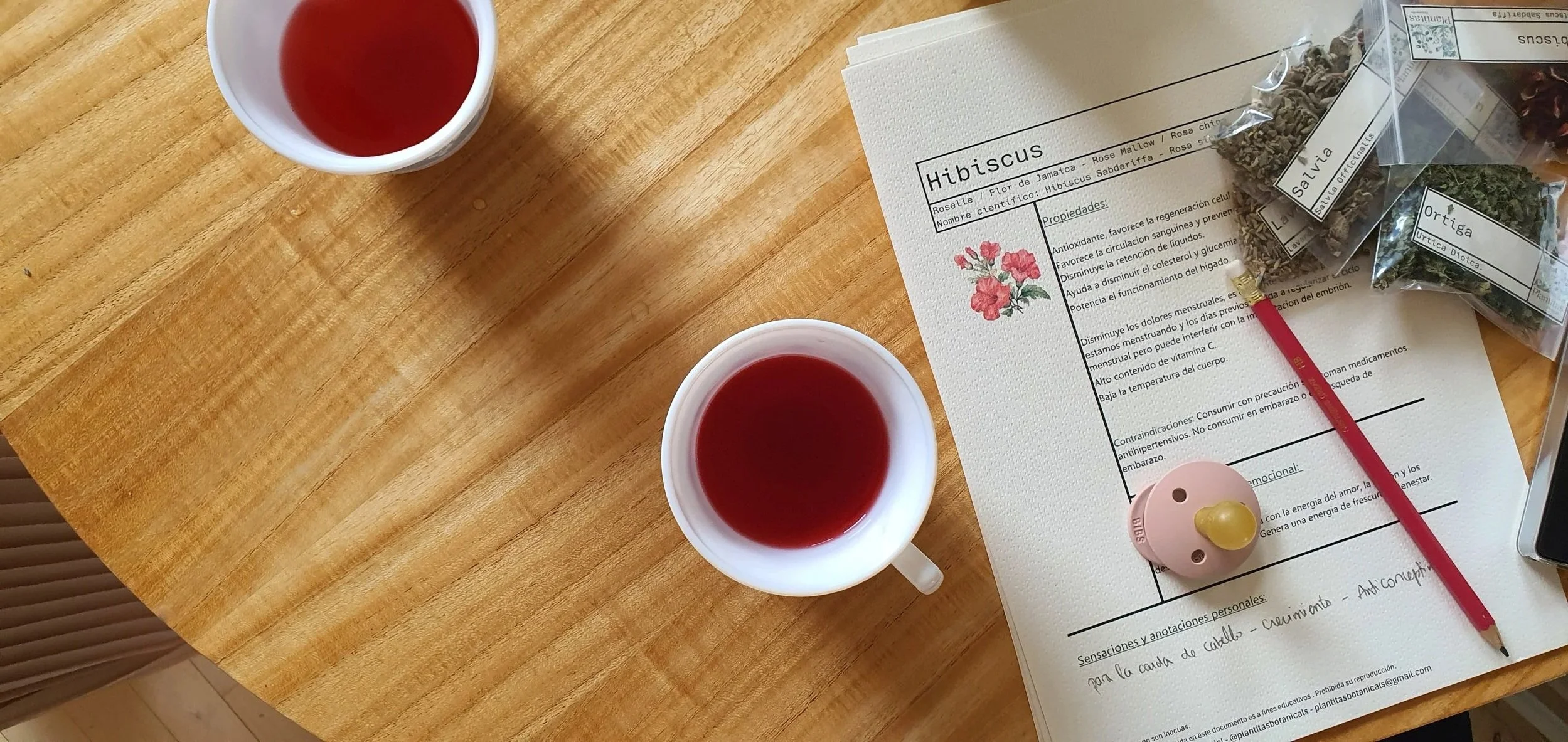 Una mesa de madera con una hoja informativa sobre la flor de hibisco, dos tazas con té de color rojo, bolsas con diferentes hierbas medicinales y una pieza de pomelo rosa.