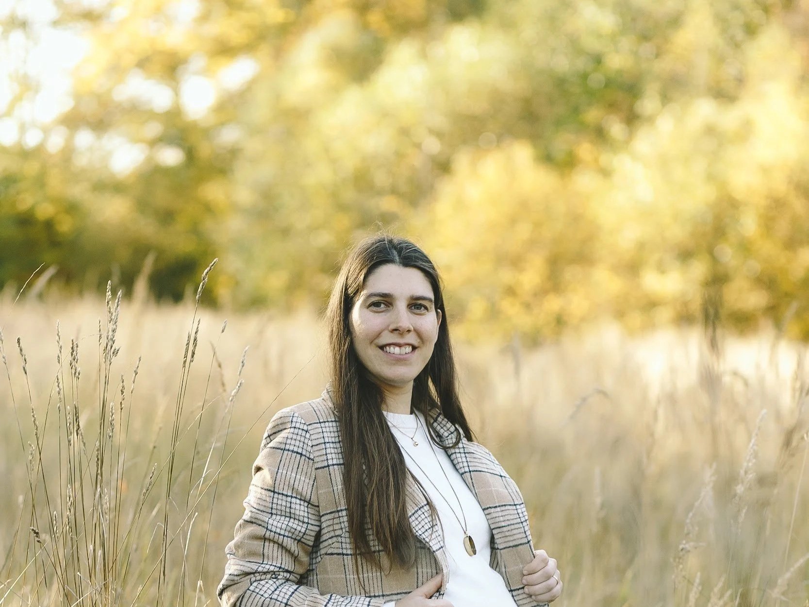 Mujer con cabello largo y oscuro en un campo con árboles de hojas verdes y amarillas en el fondo, sonriente, usando chaqueta a cuadros y blusa blanca