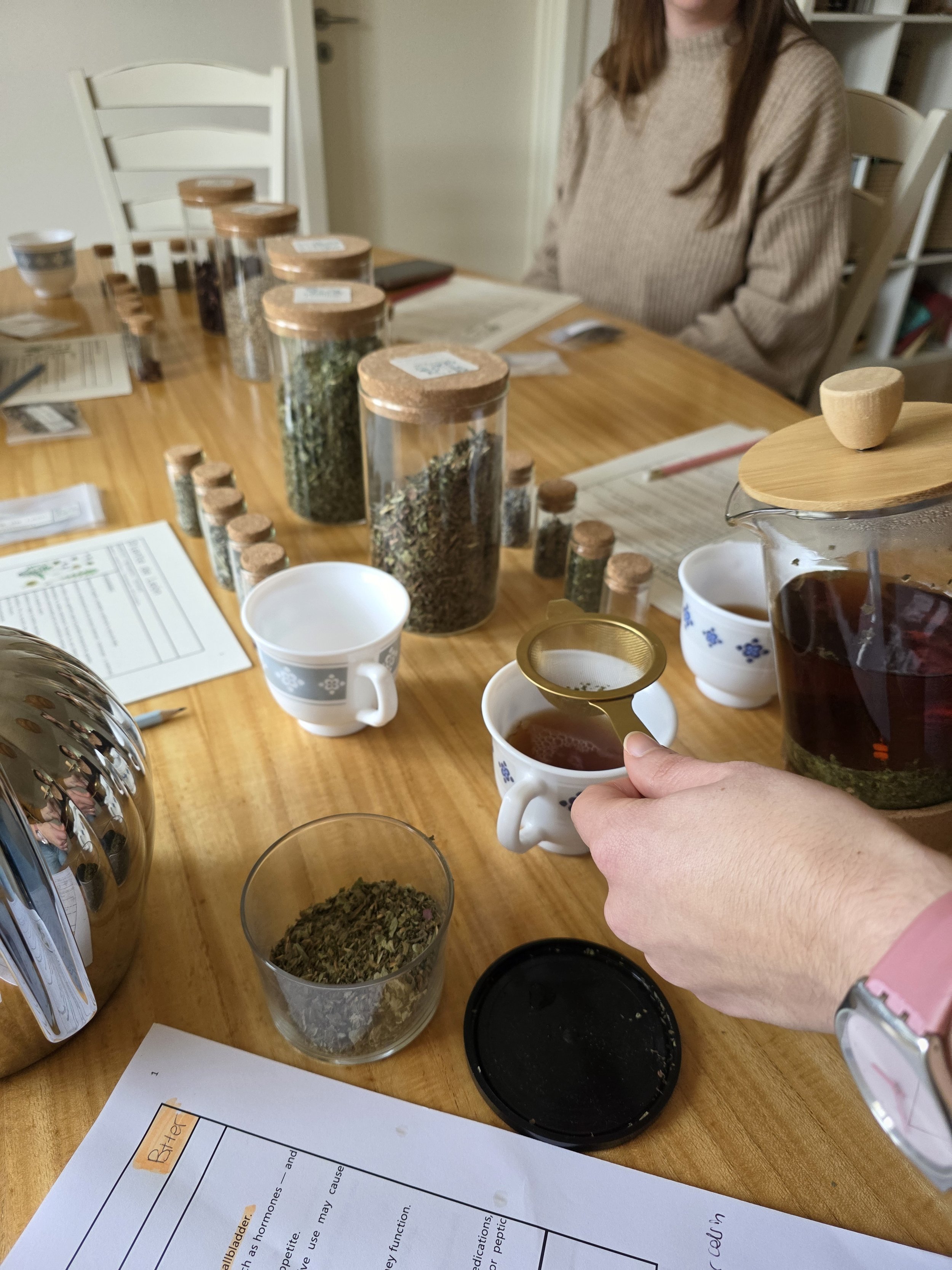 Mesa con frascos de diversas sustancias secas, tazas con té, y una pila de papeles con escritura y resaltados. Una persona sostiene una taza con té en la mano. La escena sugiere una actividad de degustación o análisis.