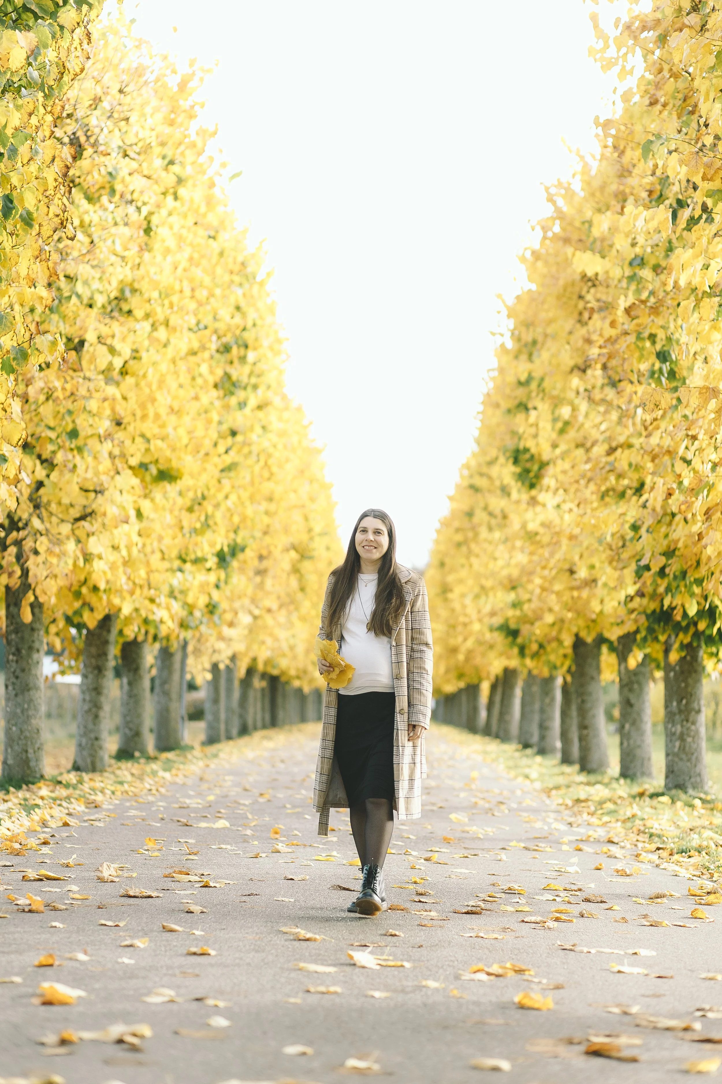 Mujer paseando por un sendero con árboles de hojas amarillas en otoño, sosteniendo hojas en una mano y sonriendo.