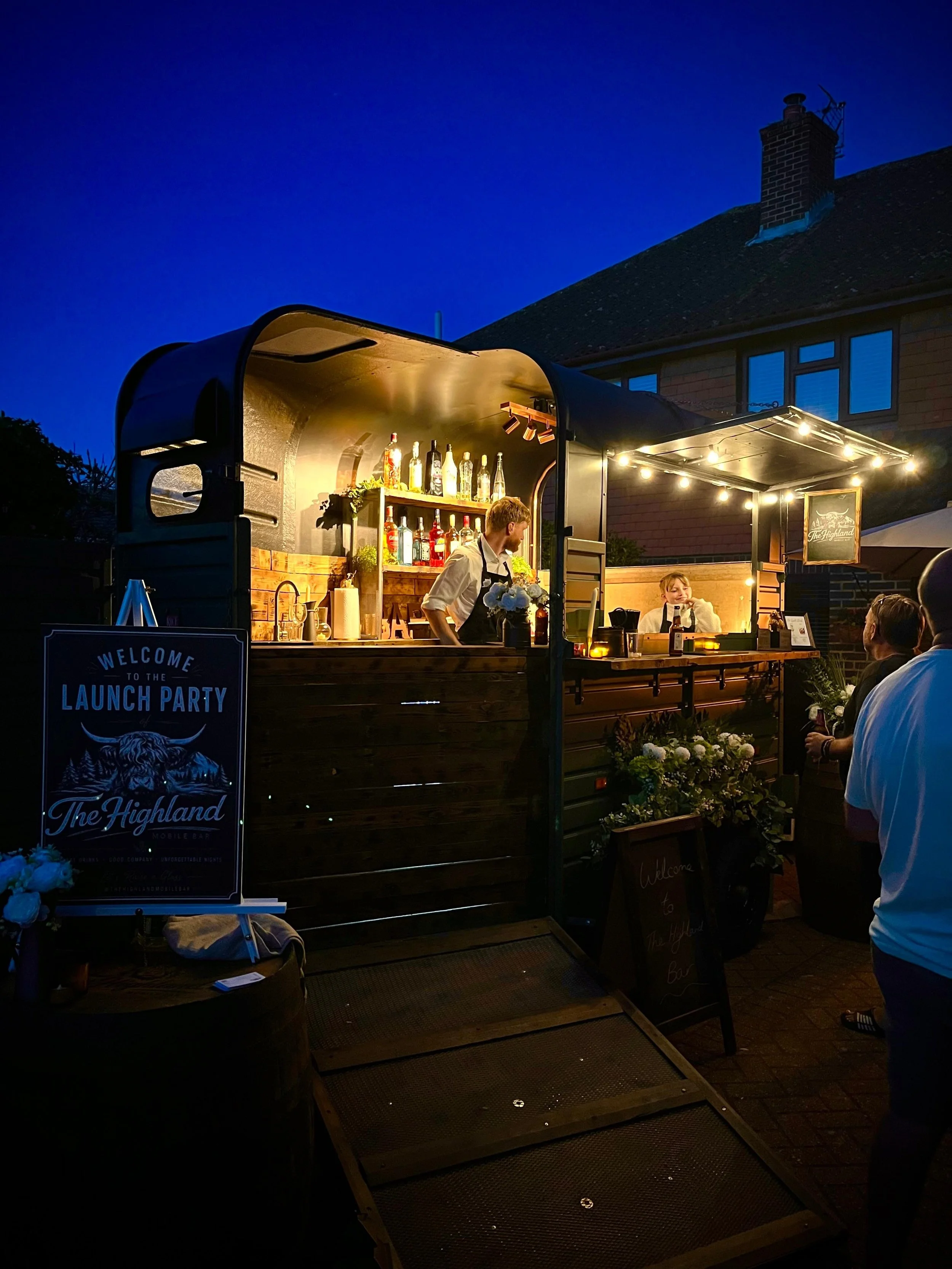 A mobile bar is set up outdoors at night, with staff serving drinks to patrons. The bar has shelves with bottles, a sign that says 'Welcome to the Highland Launch Party,' and string lights illuminating the area.