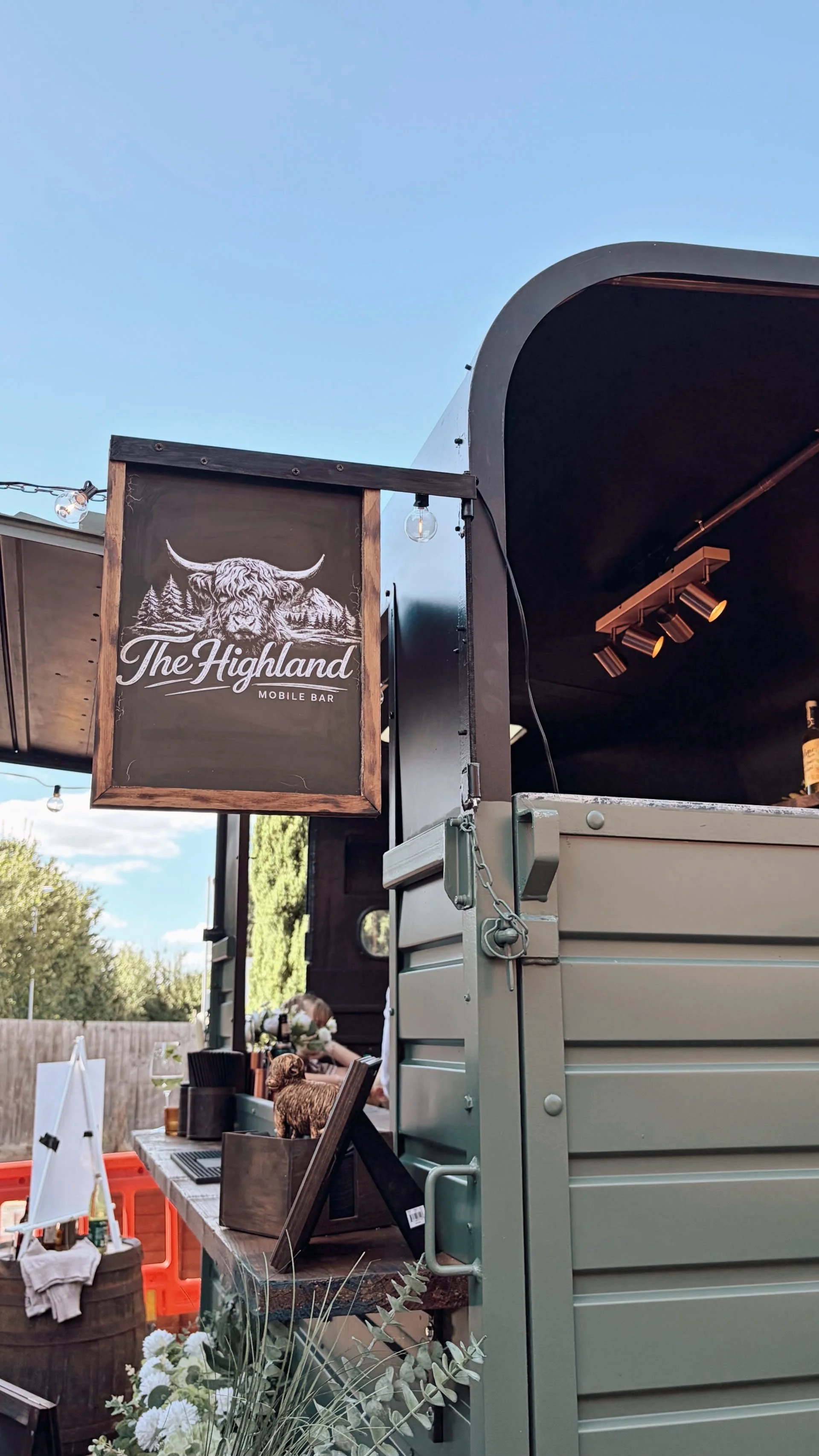 A mobile bar called The Highland with a black sign featuring a mountain and highland cow logo, set up outdoors with string lights and wooden furniture.