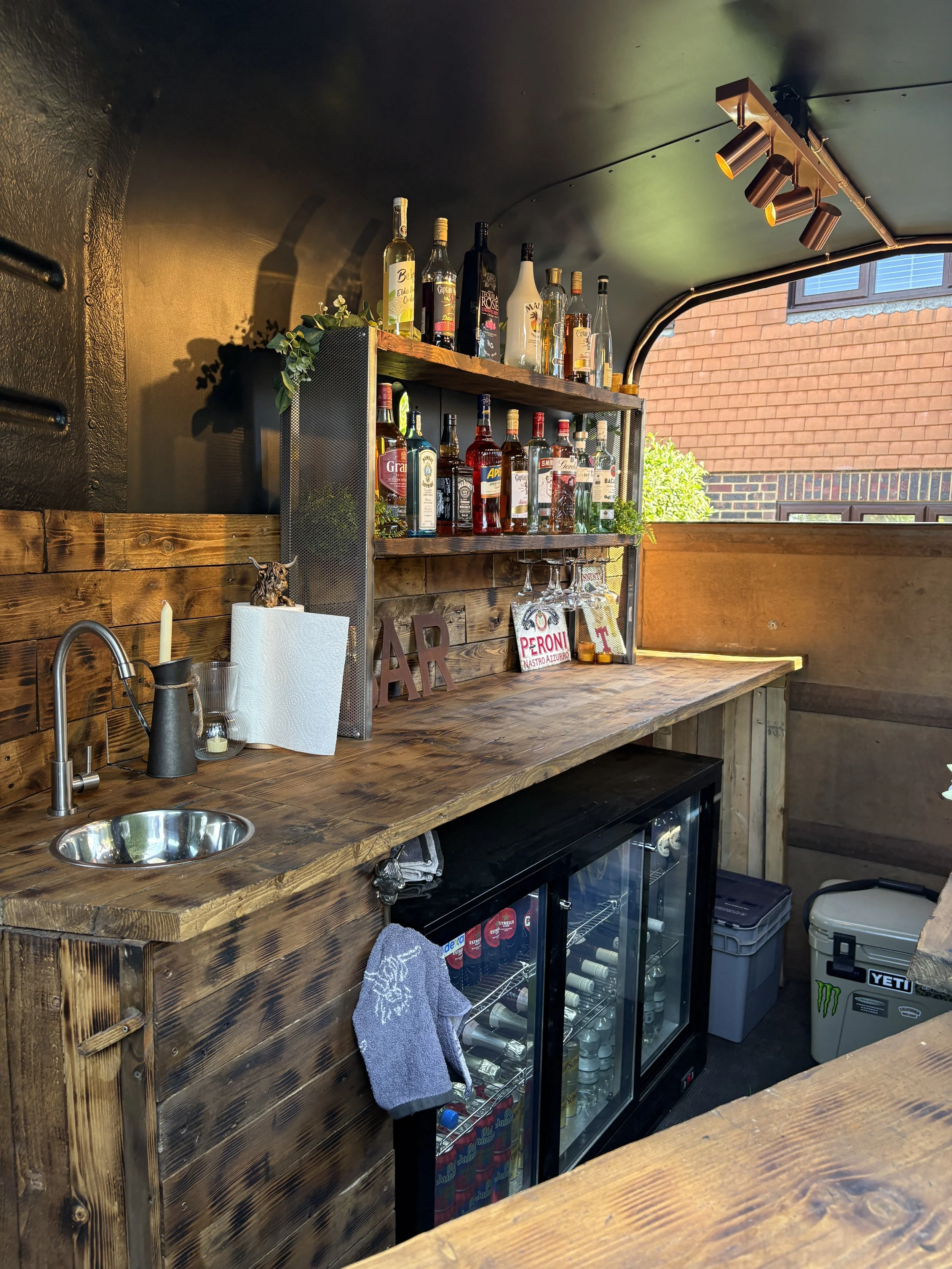 Bar area with a wooden counter, shelves stocked with various liquor bottles, a small sink, a cocktail napkin holder, and a glass-door beverage fridge underneath. There are decorative plants and a small sculpture on the shelves, and the background shows a brick wall and windows.