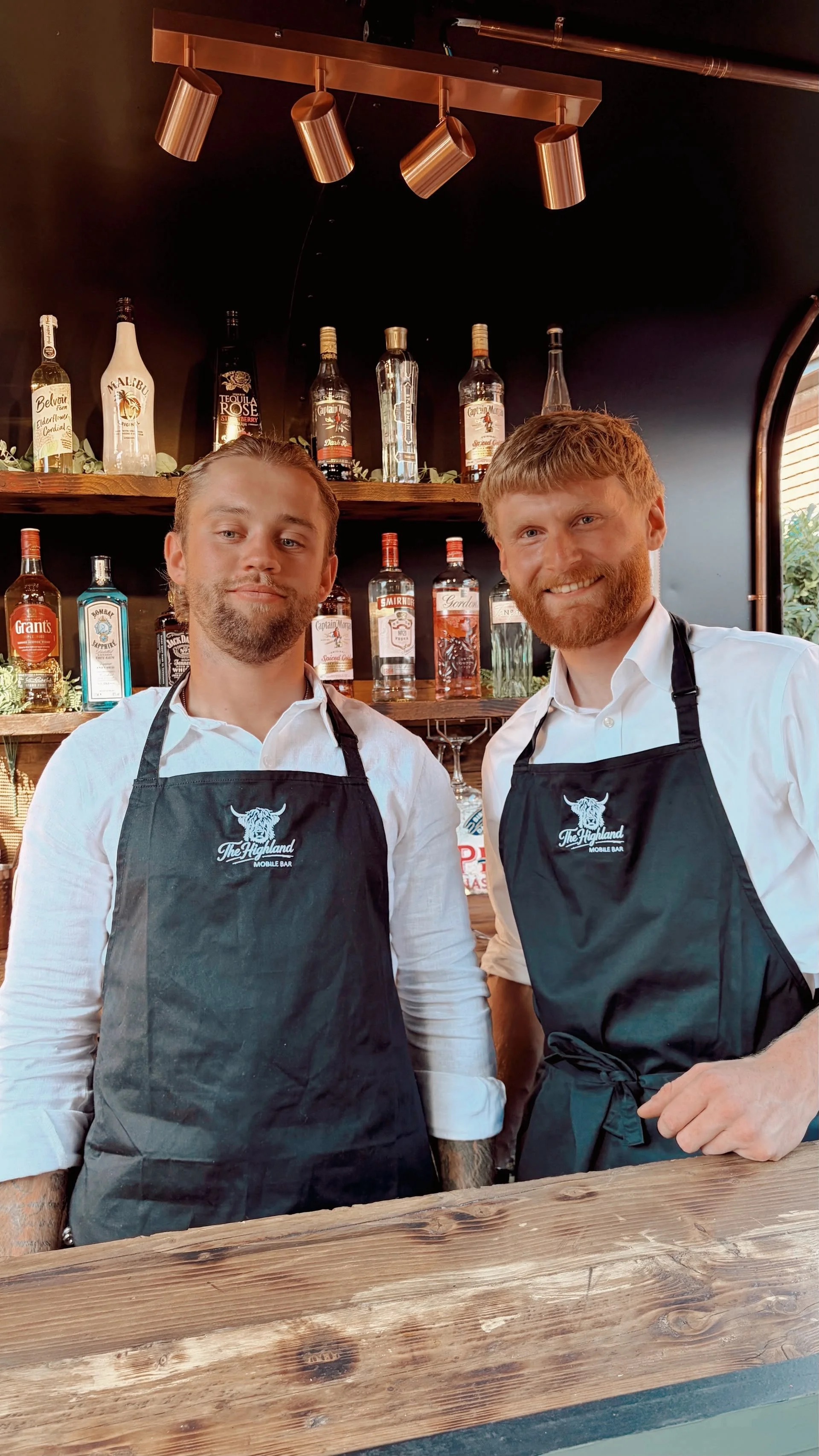 Two men wearing white shirts and black aprons with a logo, standing behind a wooden bar with bottles of alcohol on shelves behind them, inside a bar or restaurant.