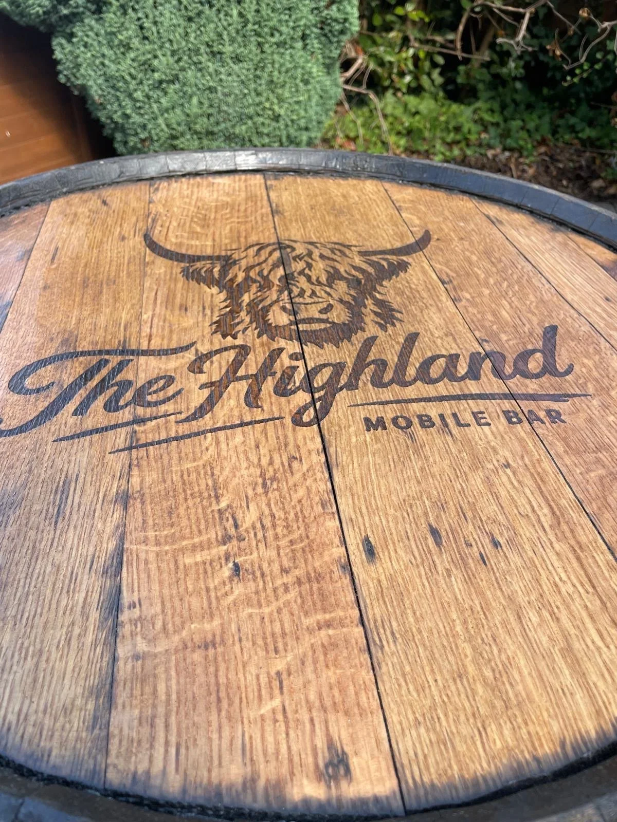 Close-up of a circular wooden table with a black border around the edge, displaying the logo for The Highland Mobile Bar, featuring a sketch of a Highland cow's head.