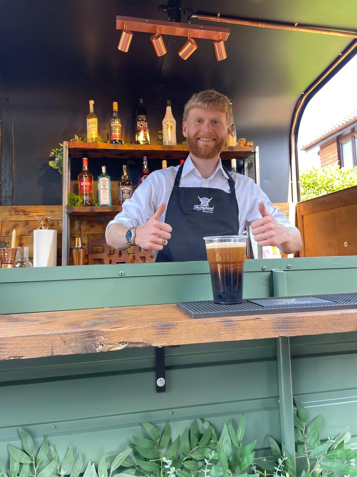 A smiling man with red hair and beard wearing a white shirt and black apron behind a bar counter. There is a drink with layered dark brown and black colors in front of him. The bar has bottles of alcohol on shelves and decor behind him, with some gre