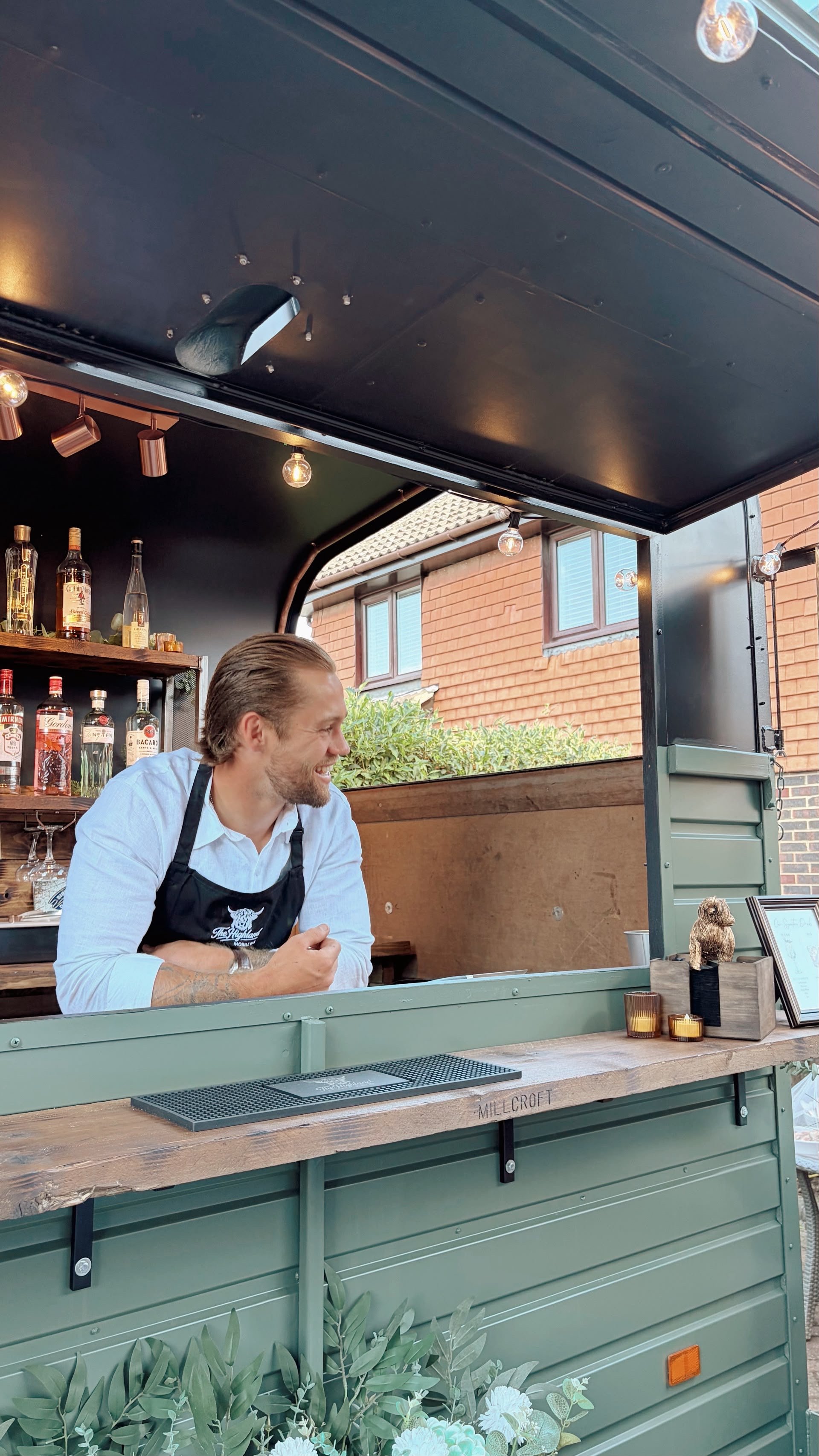A smiling man in a white shirt and black apron sitting at a counter inside a food truck, with liquor bottles on the shelf behind him. The food truck has a green exterior and is decorated with candles and a small teddy bear figure on the counter.