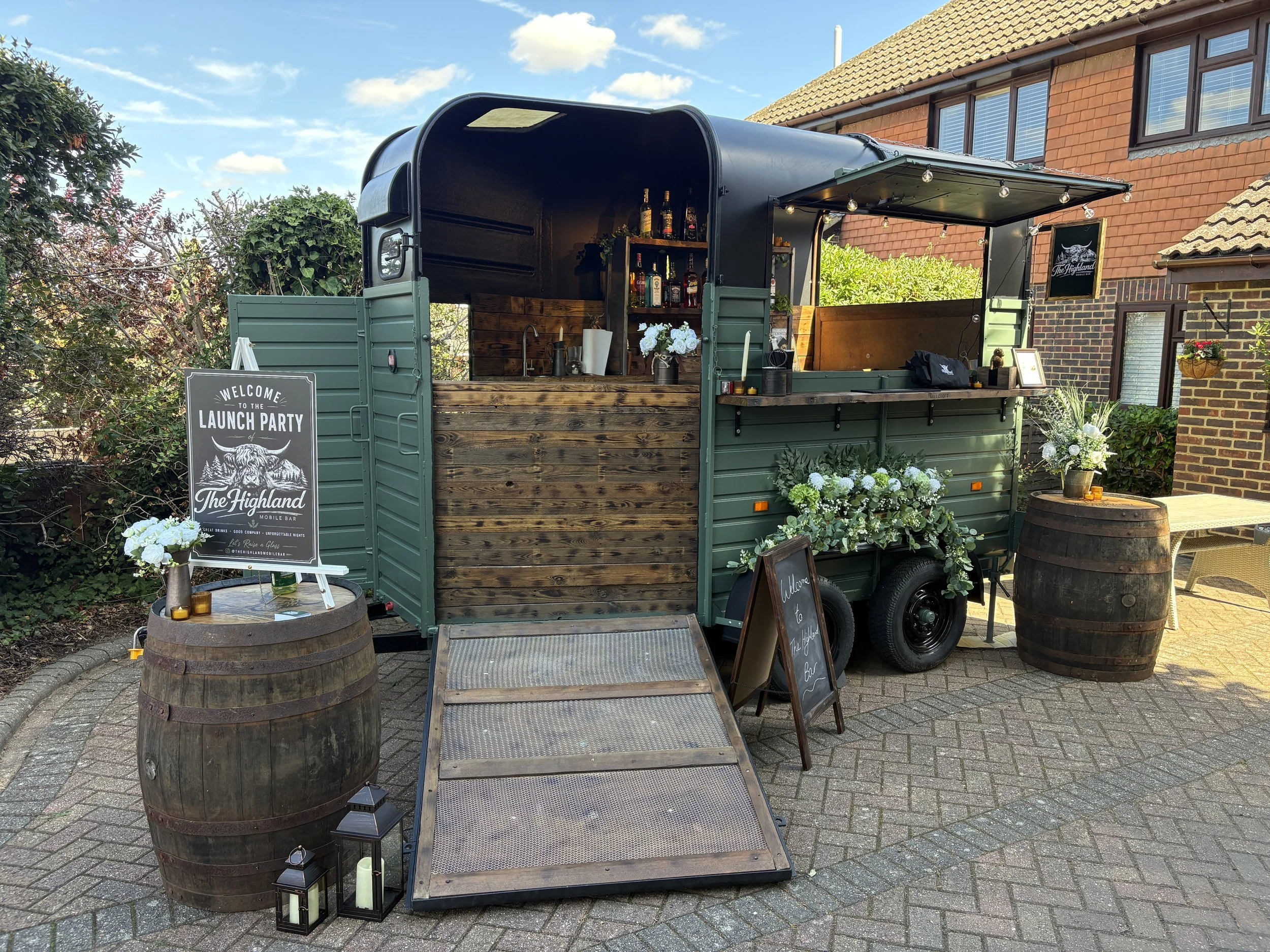 A green and wooden mobile bar set up outdoors with a sign welcoming guests to a launch party, decorated with flowers and lanterns.