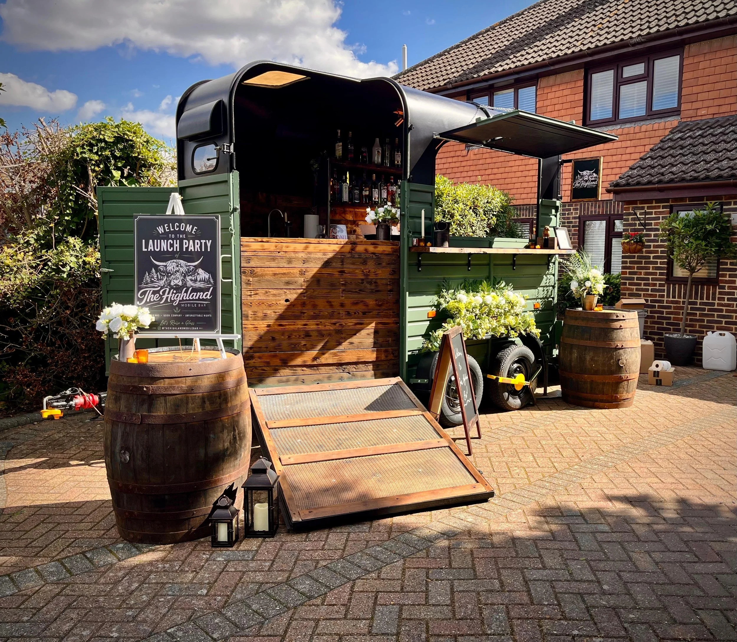 A caravan bar setup outdoors with a black and green exterior, wooden counter, and shelves with bottles, located in a residential backyard with brick houses, plants, flowers, and decorative lanterns.