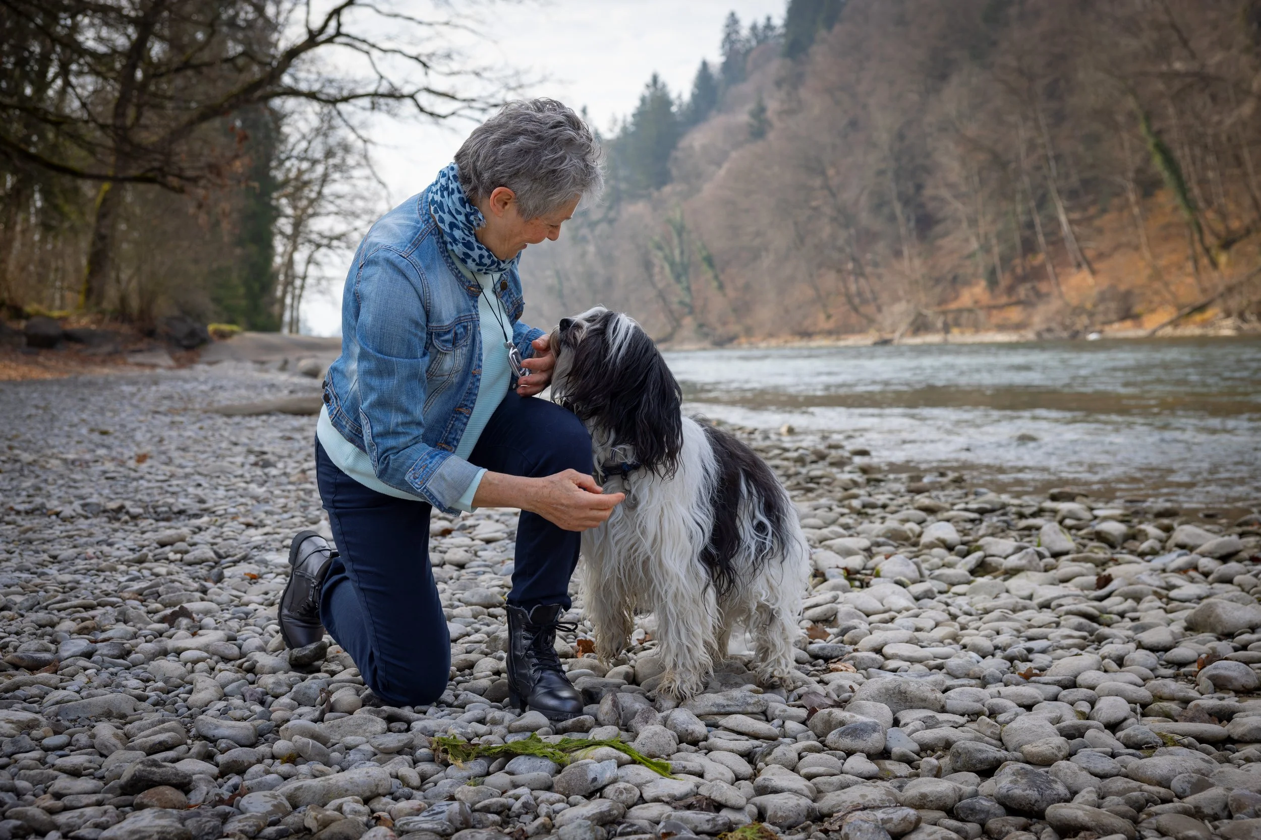 Karin Krebs kniet am Flussufer und hält Blickkontakt mit ihrem Hund, während sie ihn streichelt. Im Hintergrund sind Bäume, Felsen und Wasser zu sehen, es ist ein bewölkter Tag in einer natürlichen Umgebung.