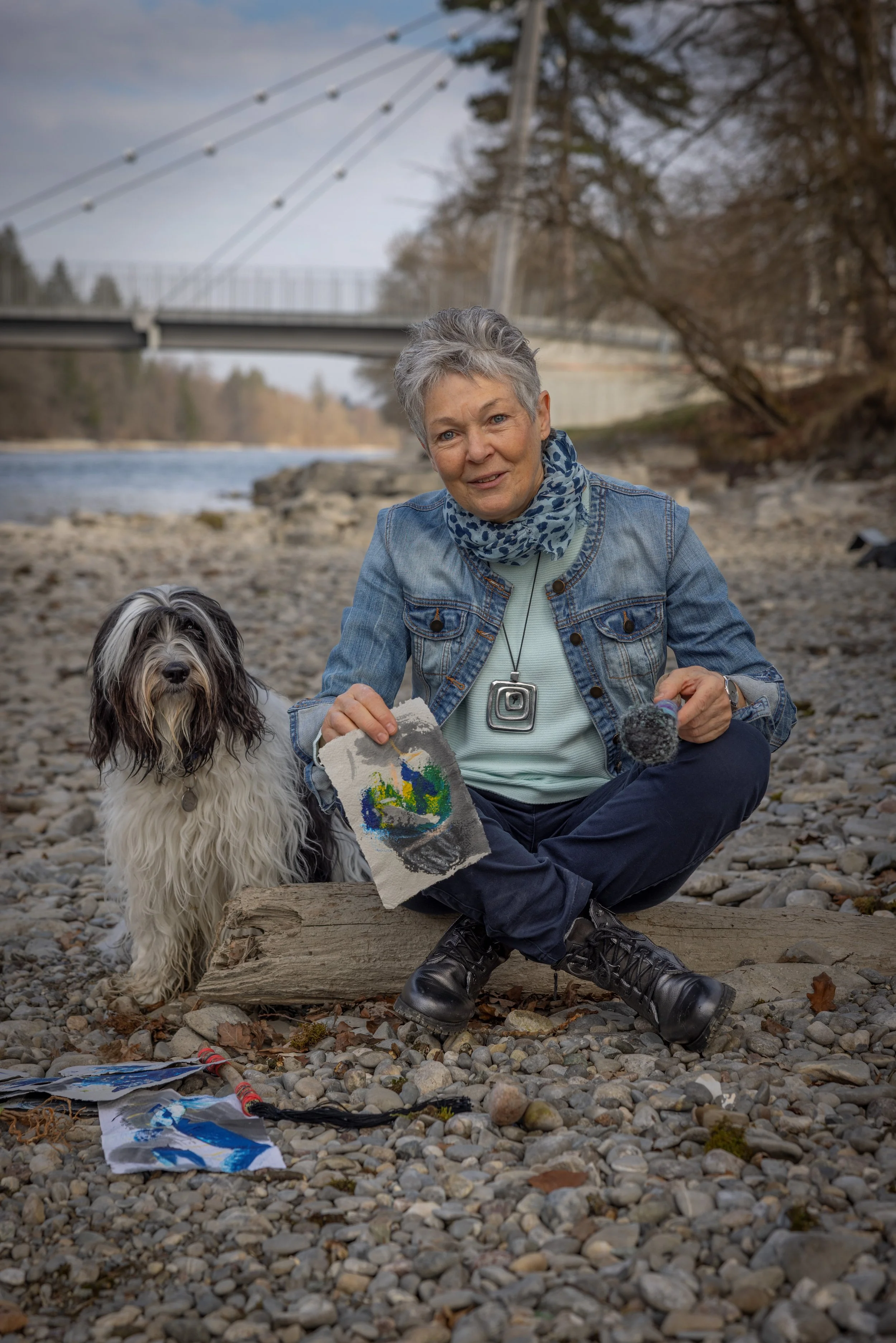 Karin Krebs, die auf einem Stein am Fluss sitzt, hält ein Stück Stoff mit einem farbenfrohen Muster, während ihr Hund neben ihr sitzt. Im Hintergrund sieht man eine Brücke und Bäumer.