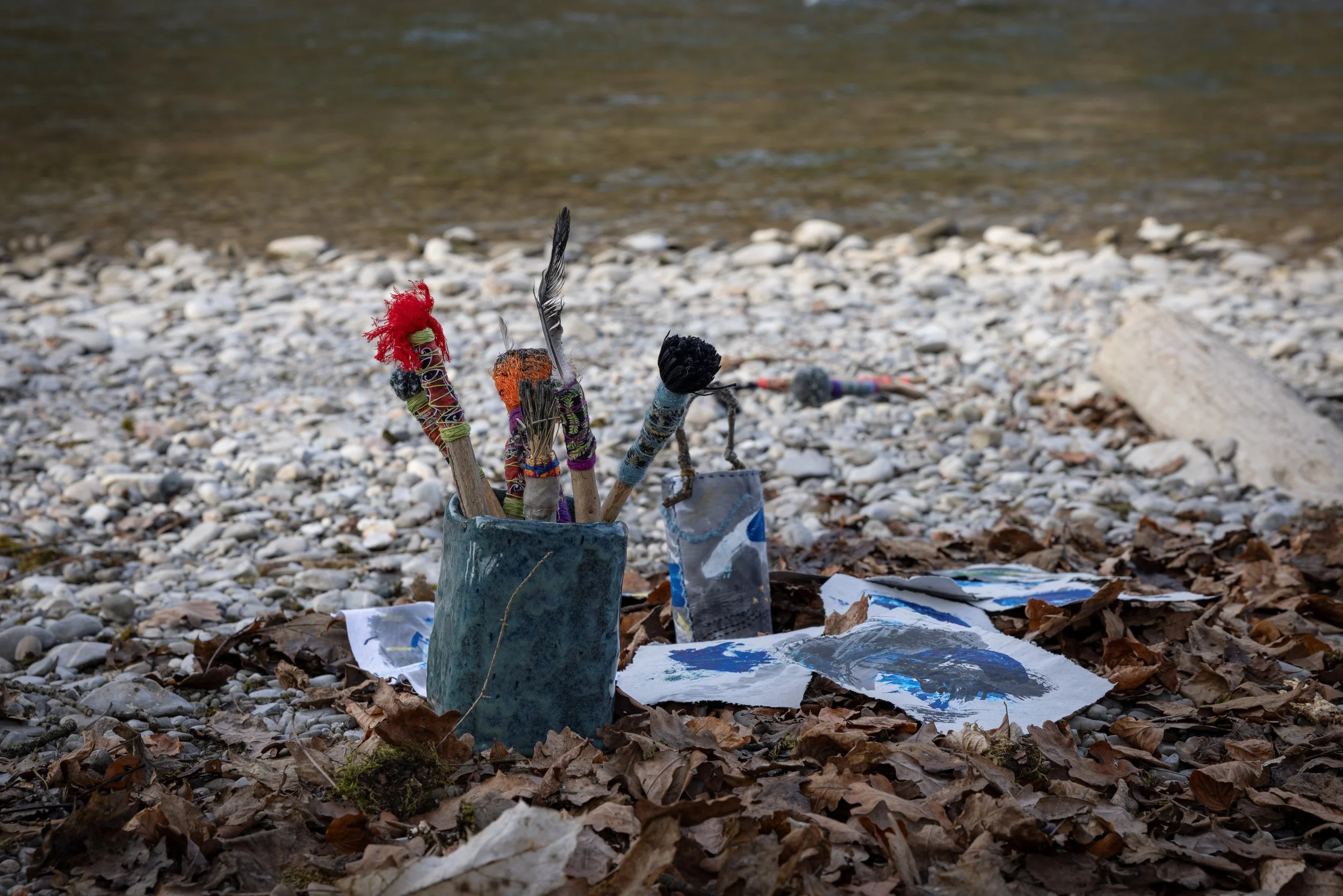 Auf einem steinigen Strand liegen bemalte Steine und Papier mit bunten Mustern, im Hintergrund das Wasser eines Flusses.