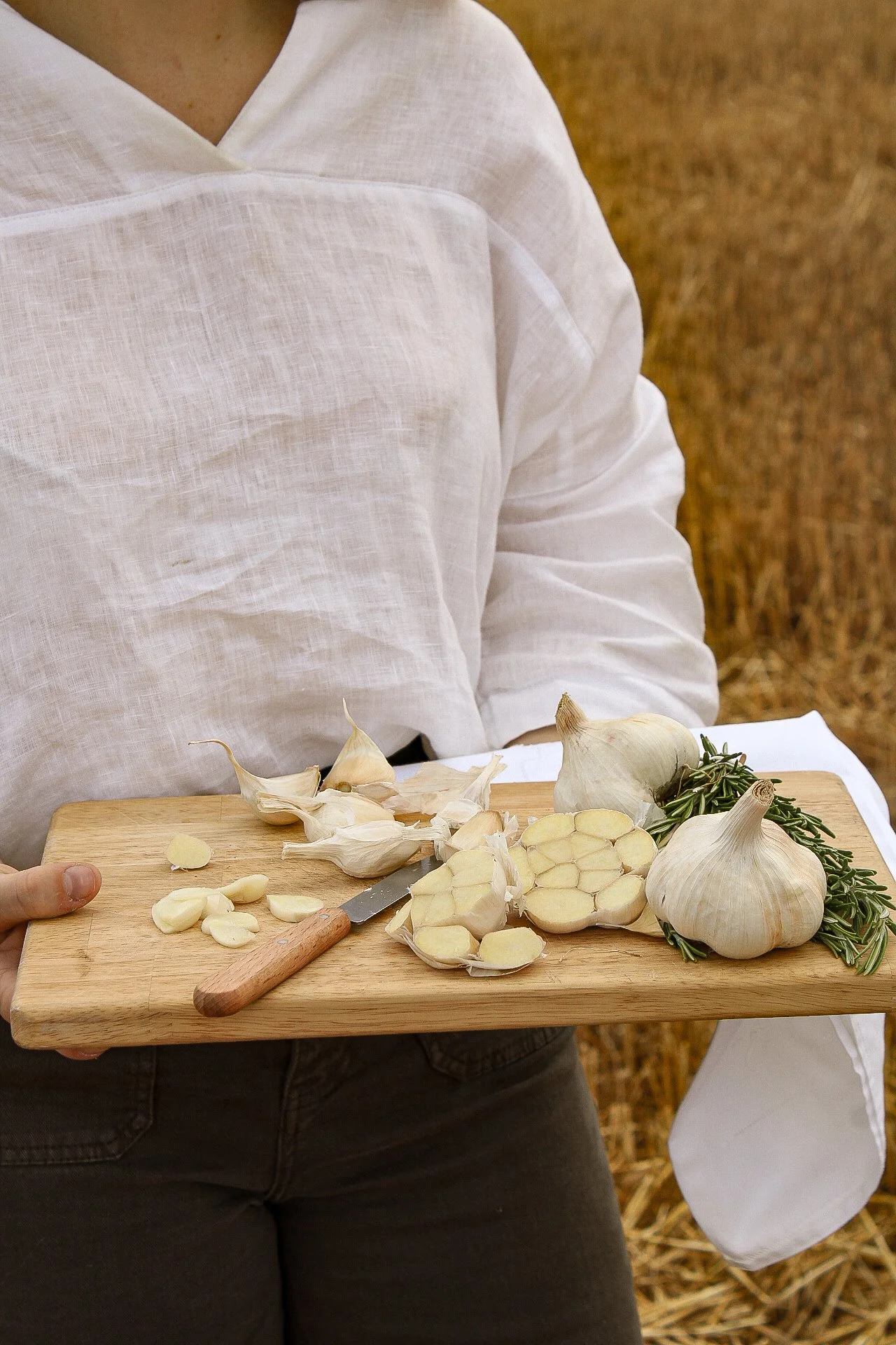 Personne portant une chemise blanche tenant une planche à découper avec de l'ail, de l'ail émincé et des herbes, dans un champ de blé.
