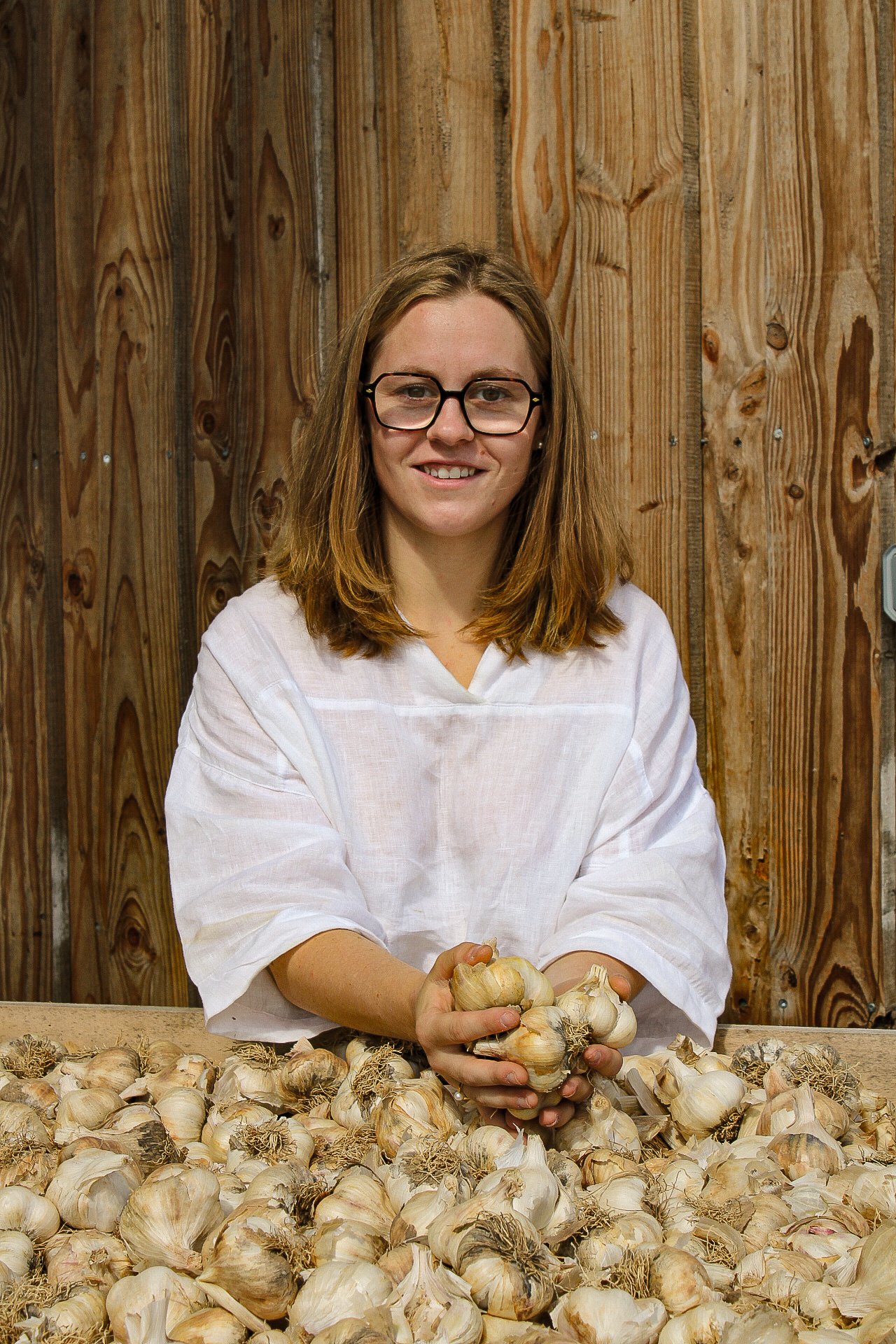 Une femme avec des lunettes, vêtue d'une blouse blanche, tient une tête d'ail dans ses mains, entourée d'ail sur une table en bois contre un panneau de bois au fond.