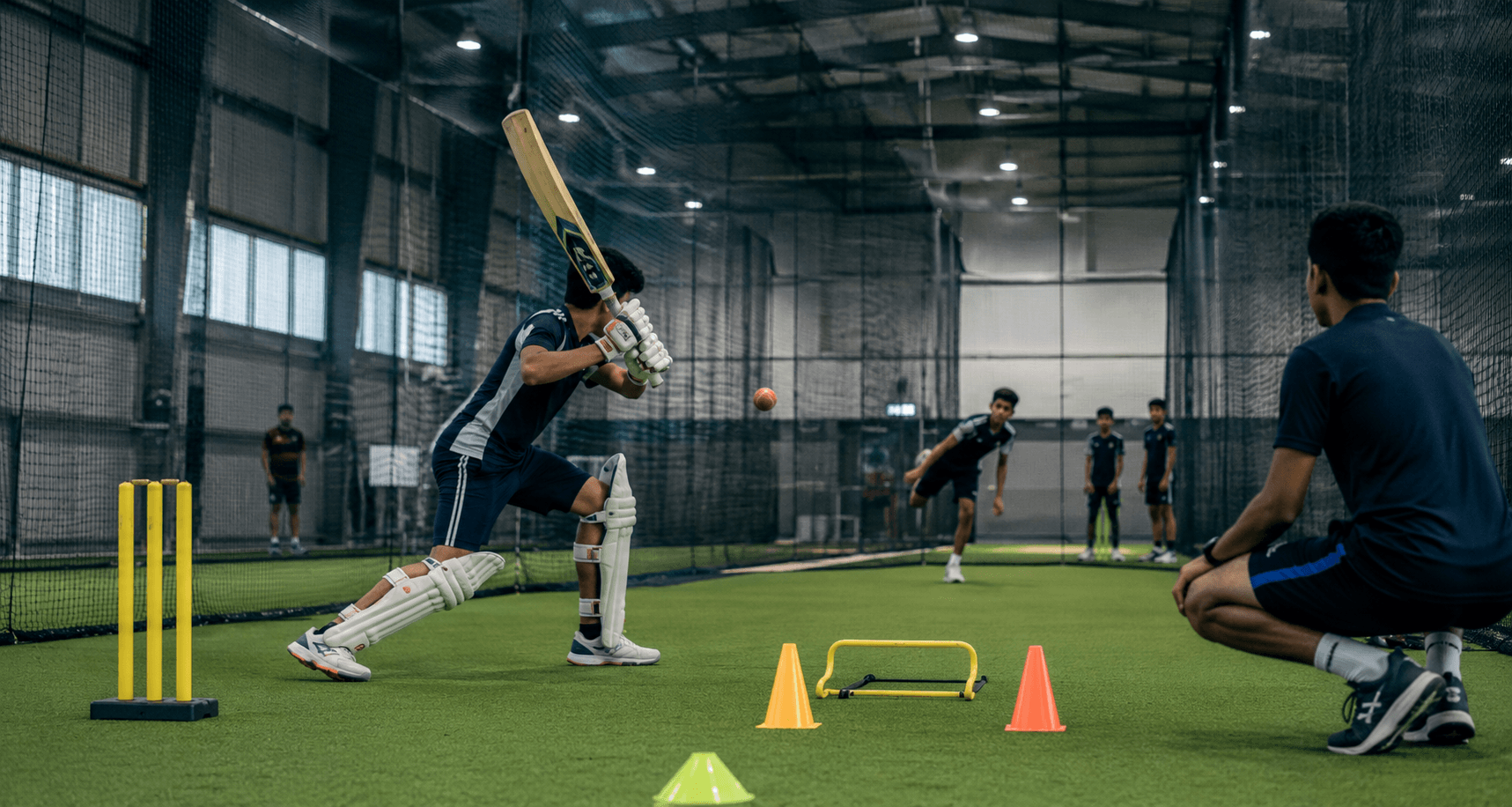 Cricket practice session inside a netted indoor facility. A young batsman with protective gear swings a cricket bat at a ball, while a coach kneels nearby. Several players are in the background, watching the practice.