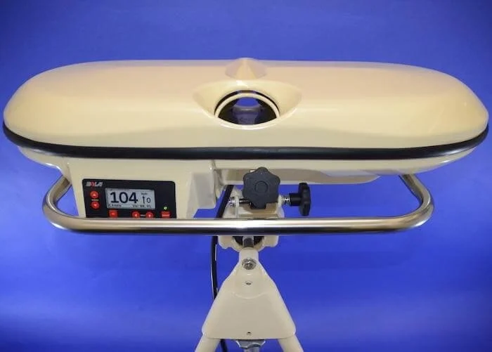 A medical device with a cream-colored oval surface, a digital display showing 104, and adjustable components on a tripod stand against a blue background.