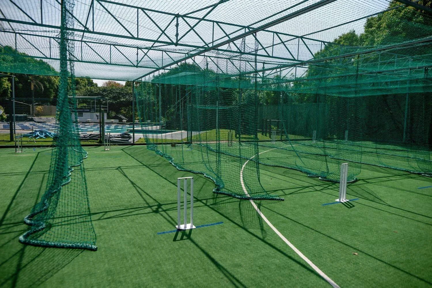 A green cricket practice net with multiple sections, set up on a synthetic turf field, with shadows cast from the netting and support structure, and trees visible in the background.