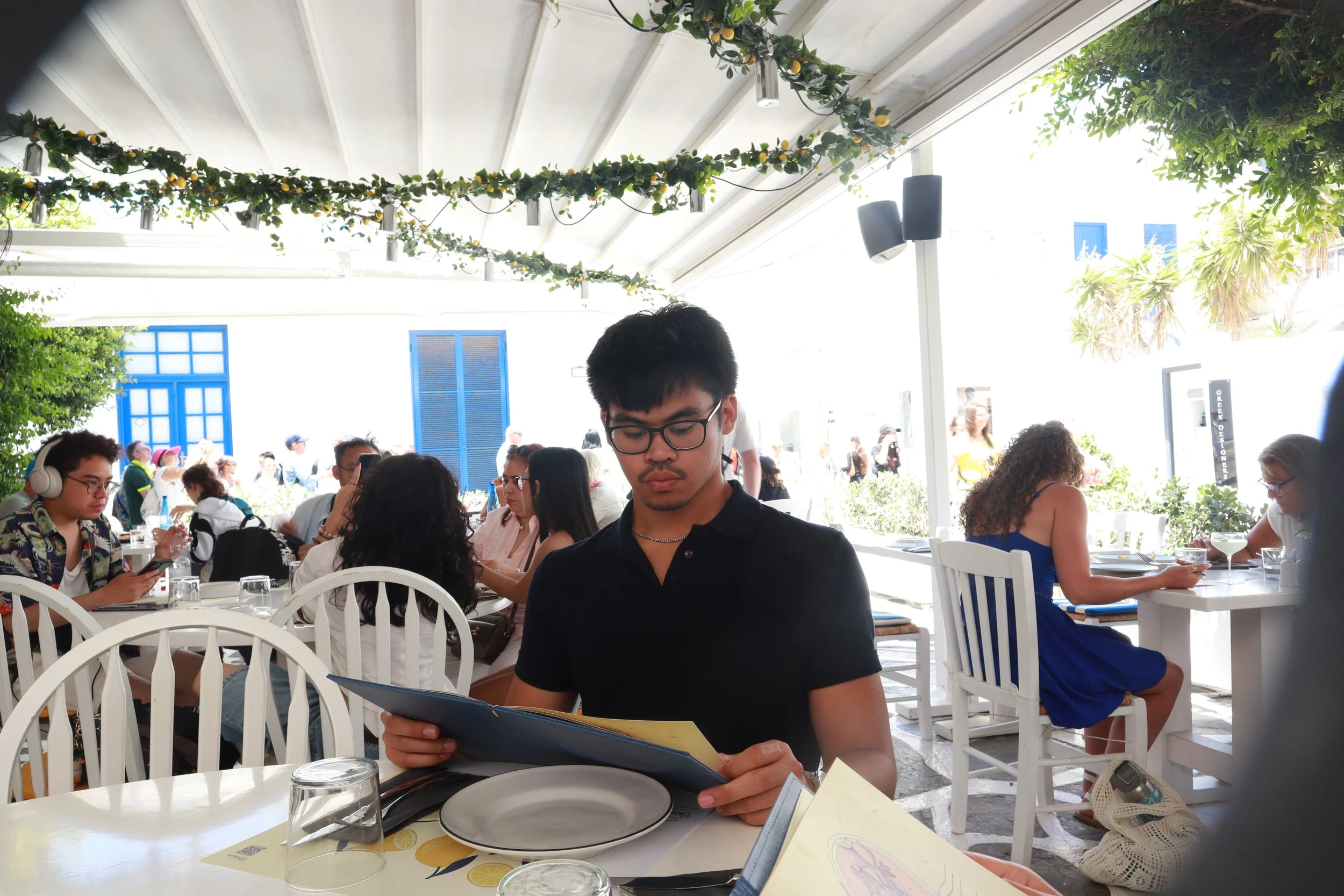 People dining at an outdoor restaurant with a white ceiling and hanging greenery, including small citrus fruits, on a sunny day.