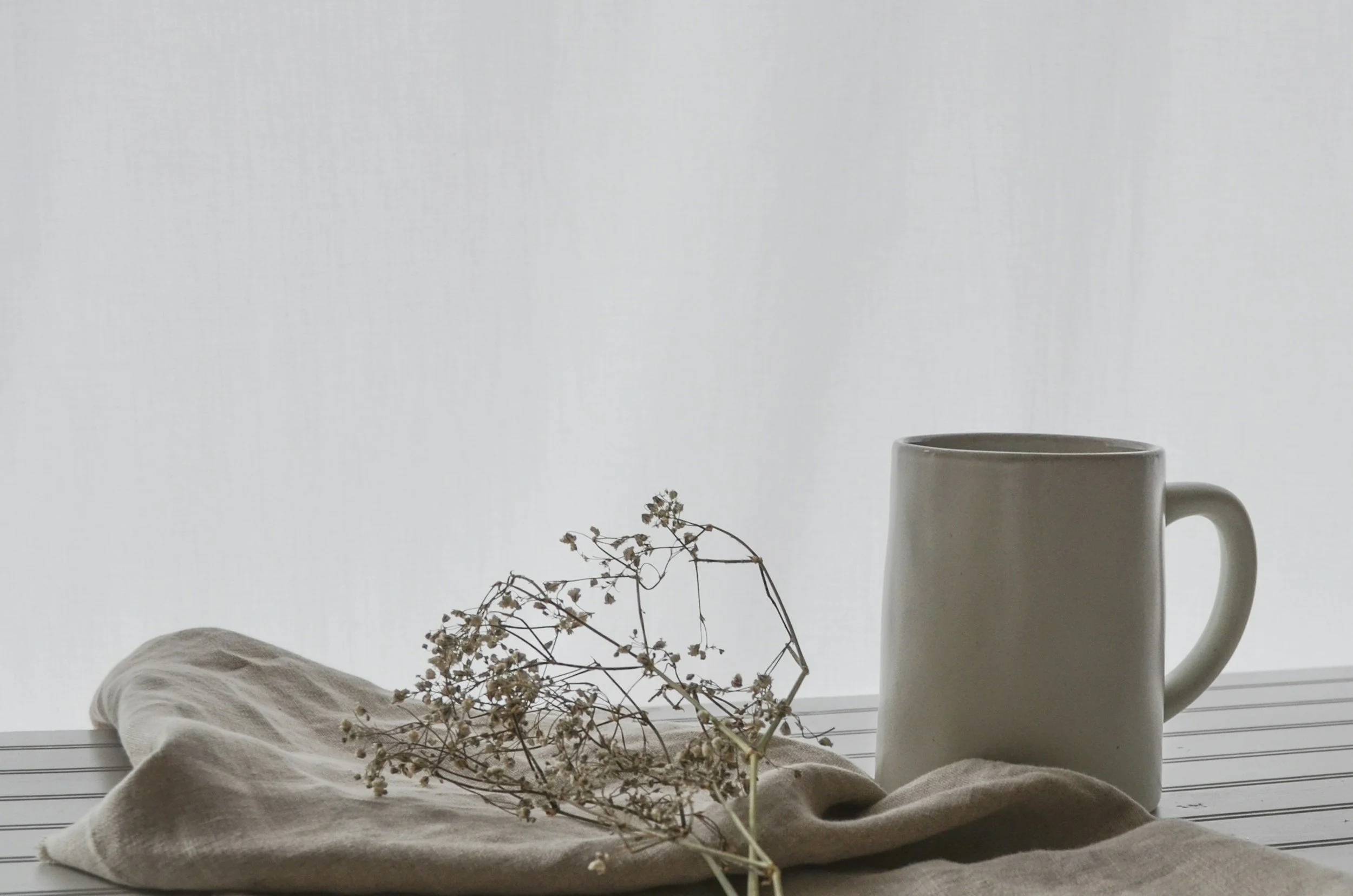 Une tasse blanche avec une anse à droite, posée sur une surface blanche. À gauche, des branches de fleurs séchées sur un tissu beige, par une fenêtre avec la lumière naturelle en arrière-plan.