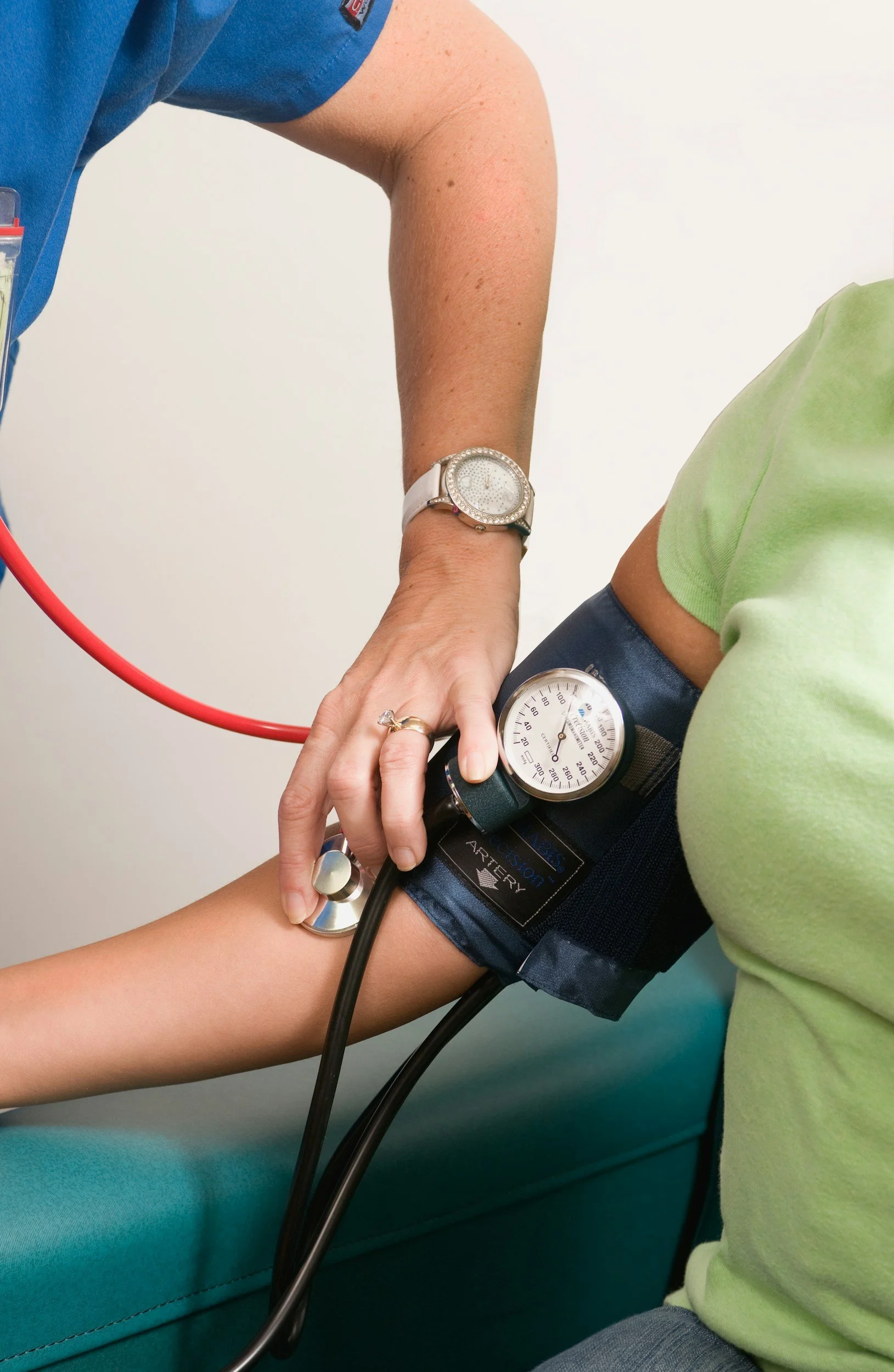 Une personne en tenue de soins médicaux prenant la pression artérielle d'un patient à l'aide d'un tensiomètre au bras, dans un environnement médical.