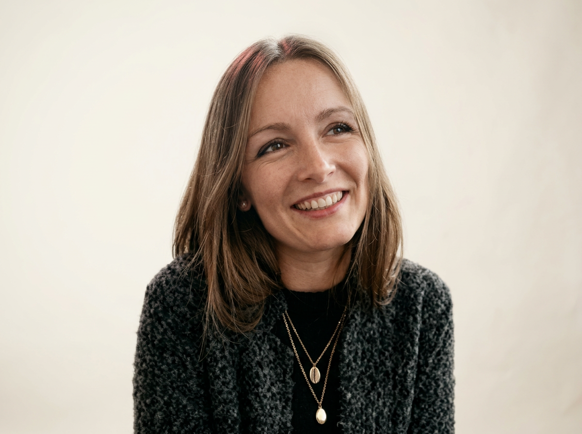 Jeune femme souriante avec des cheveux bruns, portant un pull noir et un blazer à motifs, avec deux colliers pendentifs, devant un mur clair.
