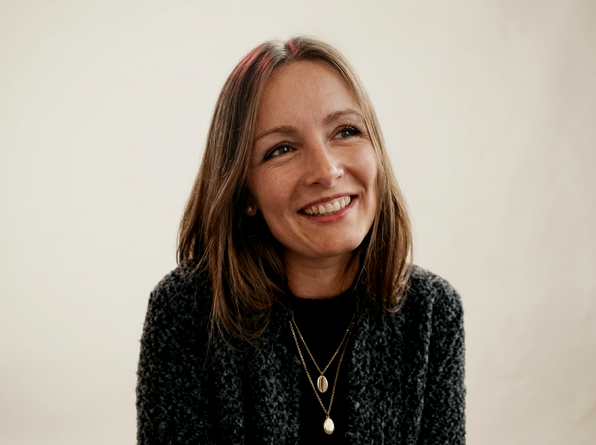Jeune femme souriante avec des cheveux bruns, portant un pull gris et plusieurs colliers, devant un fond clair.