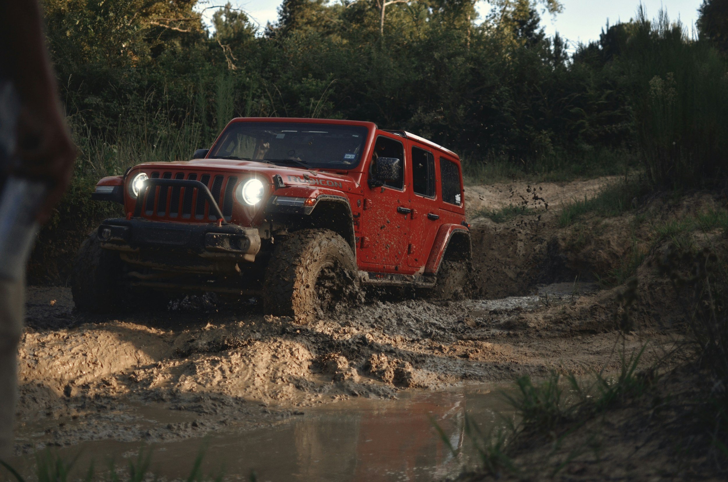A red Jeep Rubicon off-road vehicle driving through muddy terrain with water splashing, surrounded by green trees and bushes in a forested area.