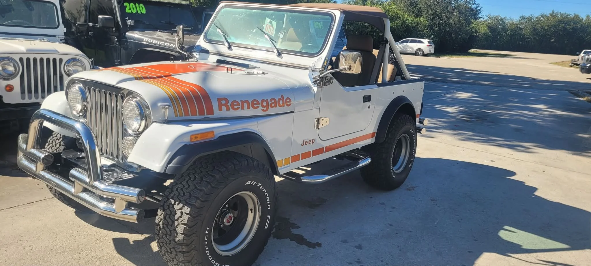 A vintage white Jeep with orange and yellow racing stripes and the word 'Renegade' on the side. It has large off-road tires, a chrome front bumper, and a roll cage. The Jeep is parked outdoors in a lot with other vehicles and trees in the background, on a sunny day.
