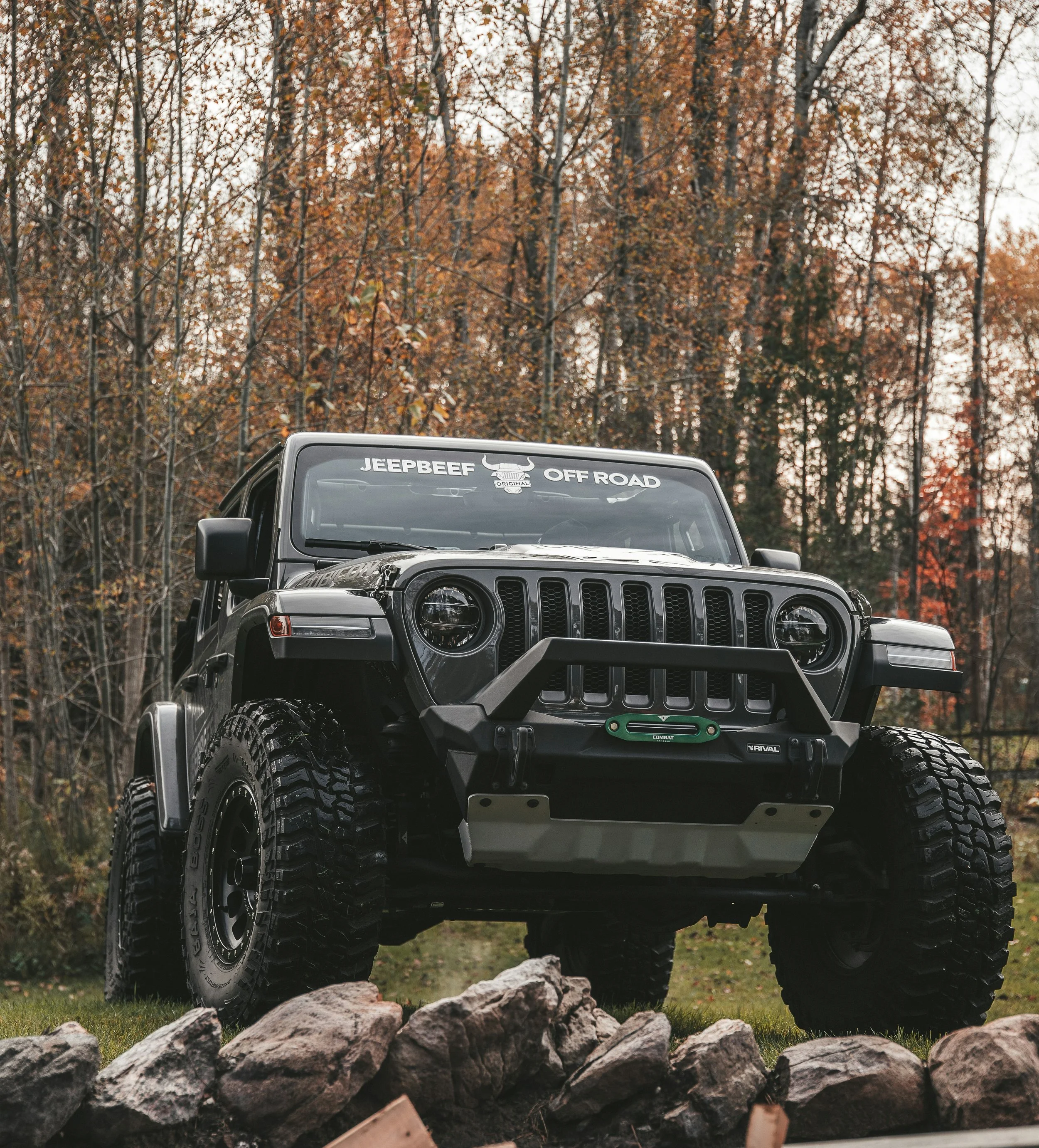The front of a black Jeep with off-road tires parked outdoors surrounded by rocks and trees with autumn leaves.