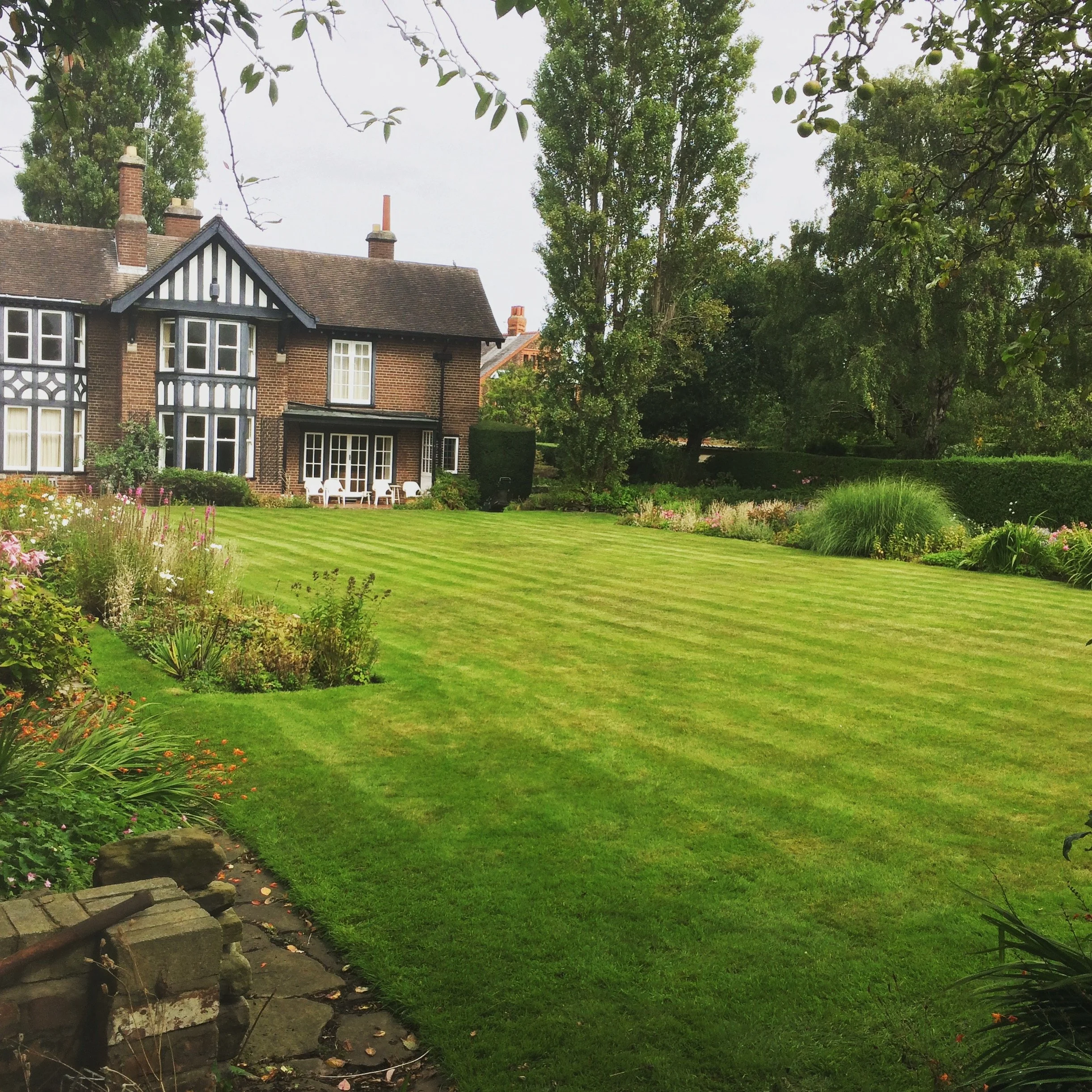 A large brick house with multiple chimneys and windows, surrounded by a well-maintained garden with flowers, trees, and a grassy lawn, with a stone pathway in the foreground.