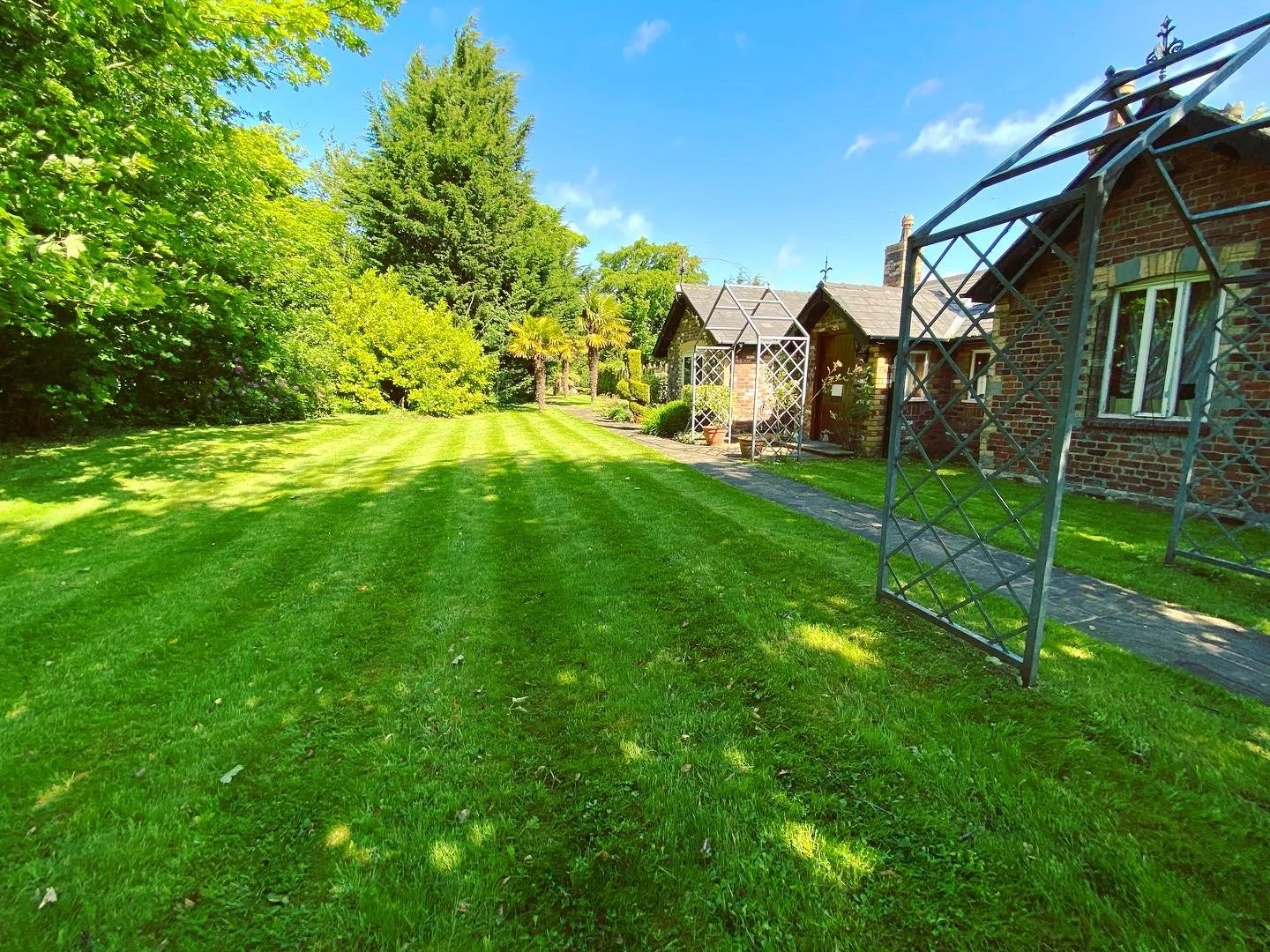A lush green backyard with well-maintained lawn, various trees, and a brick house with metal garden structures.
