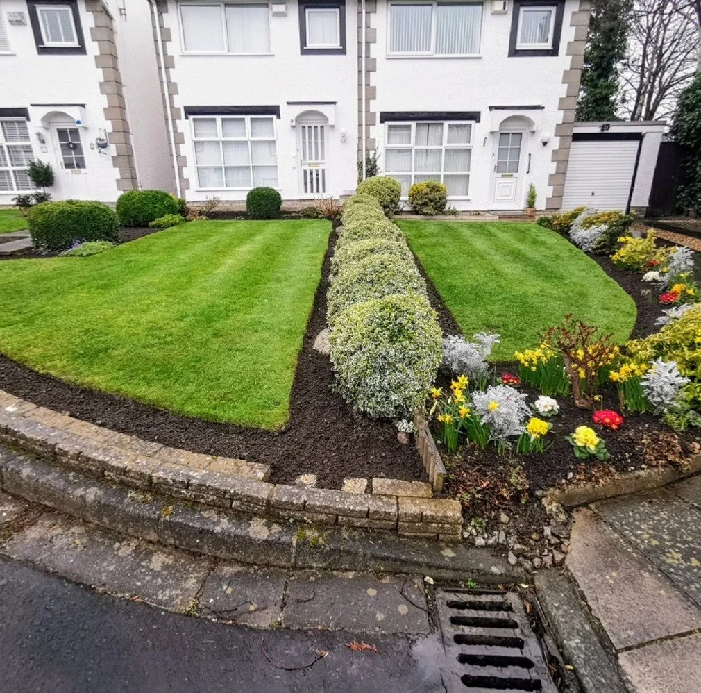 Well-maintained front yard with neatly trimmed grass, decorative bushes, and colorful flowers bordered by a small brick wall in front of a white house with multiple windows.