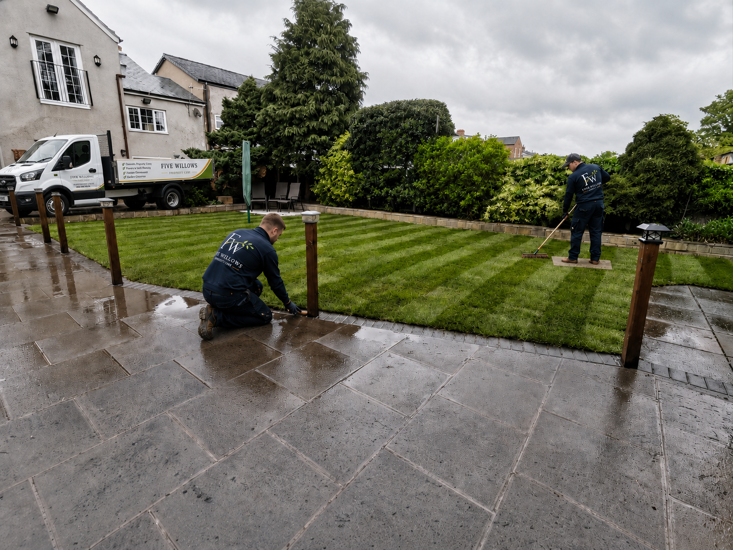 Two workers planting grass on a lawn, with a paved patio in the foreground, a truck labeled 'Five Willows' parked nearby, and residential buildings with trees in the background, all under a cloudy sky.