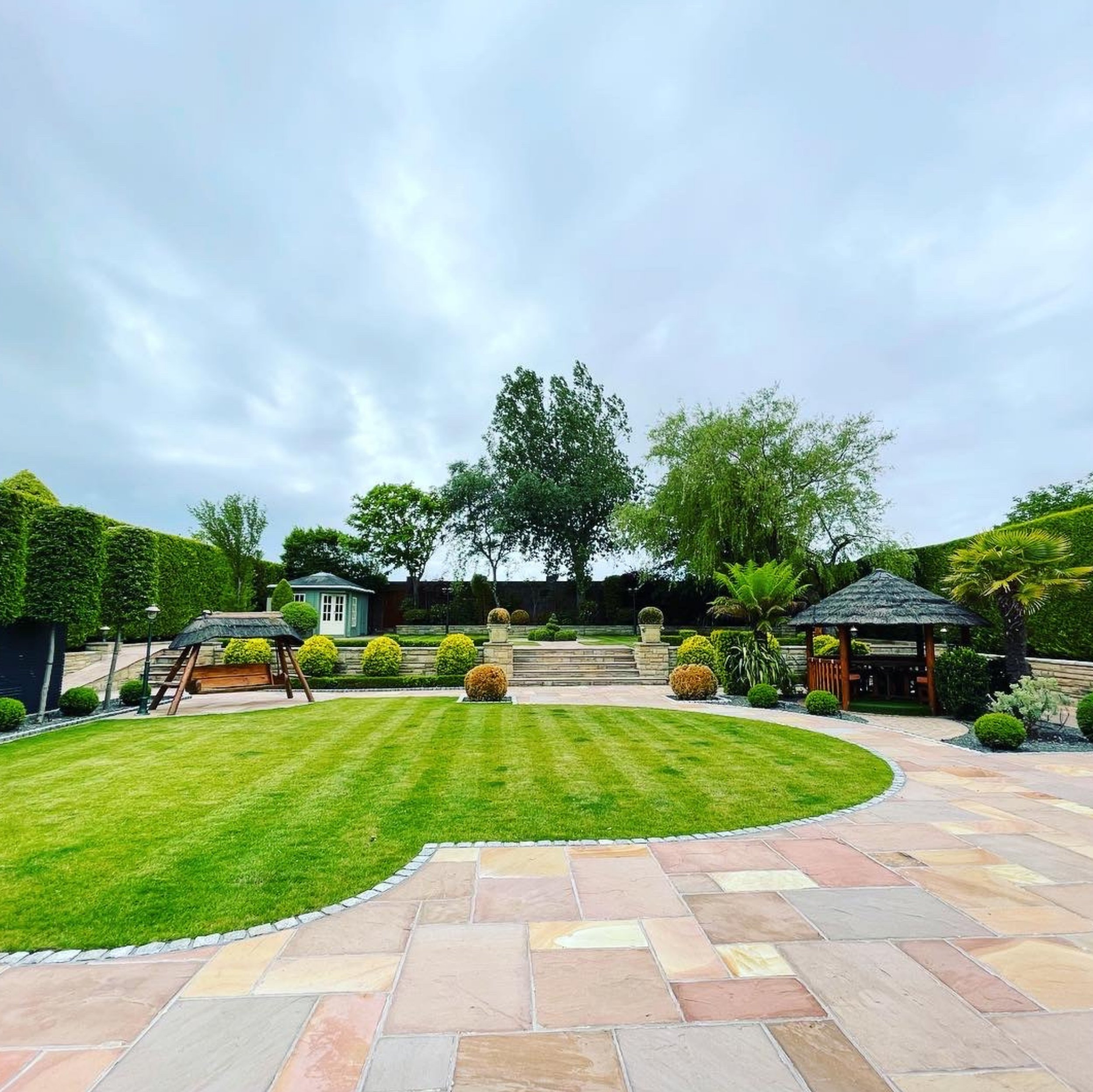 A garden with a neatly manicured lawn, a paved walkway, and various plants and trees, under a cloudy sky.
