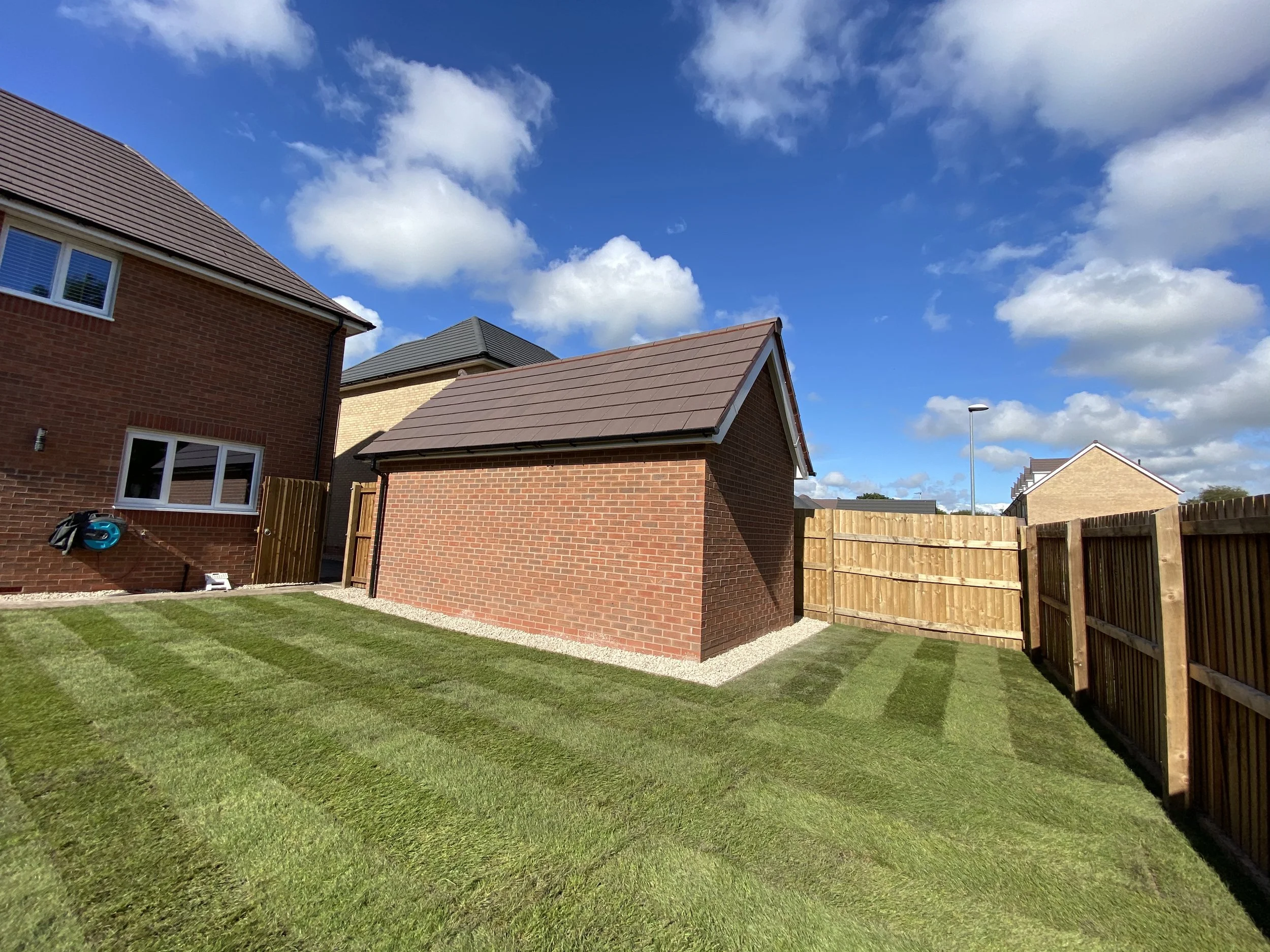 A backyard with neatly trimmed grass, a brick house, a wooden fence, and a partly cloudy sky.