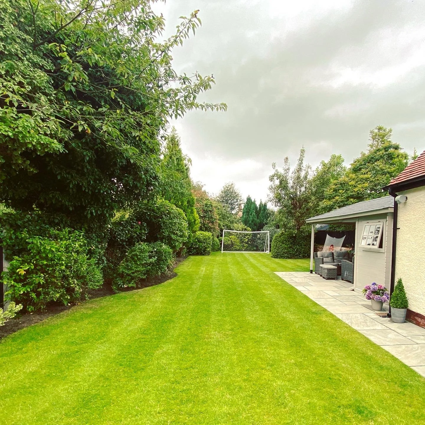 A lush green backyard with neatly mowed grass and a garden shed on the right. The shed has patio furniture and potted plants outside. In the background, a soccer goal is visible on a cloudy day.